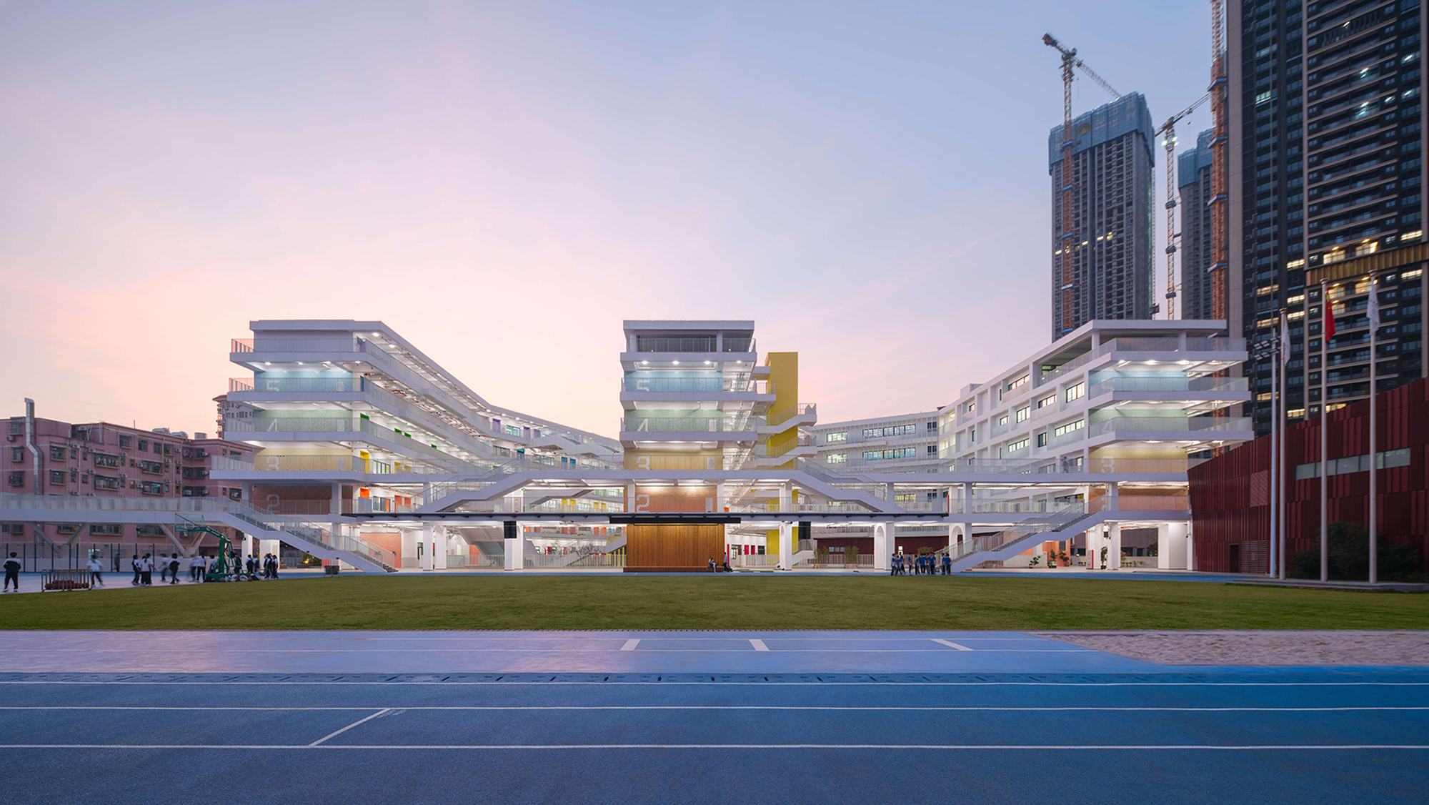Layered white facade with yellow accent tower viewed across a blue running track at dusk