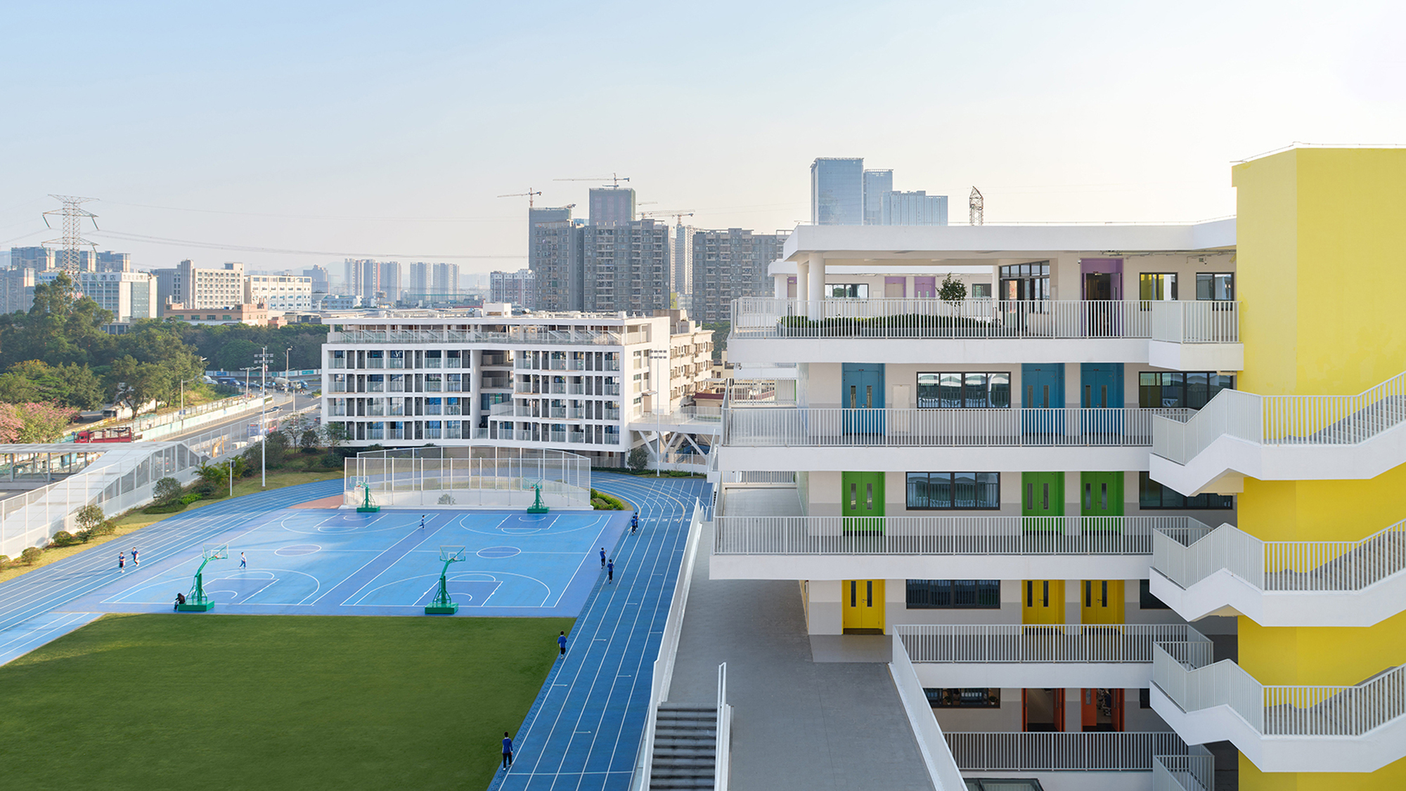 Terraced school building with colored balcony screens overlooking sports courts and distant high-rise towers
