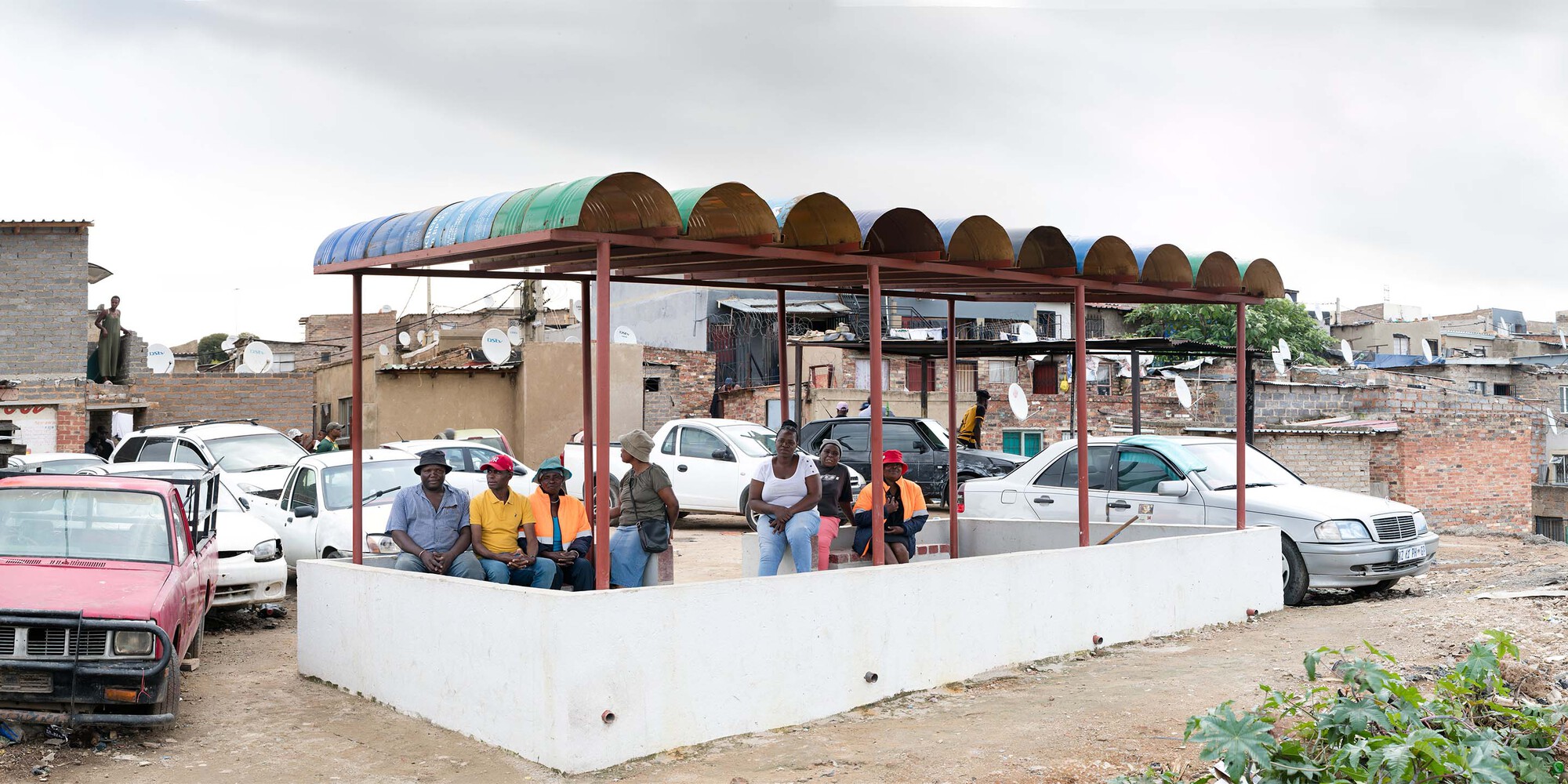 Covered community gathering platform with semicircular roof panels and people seated on white painted benches