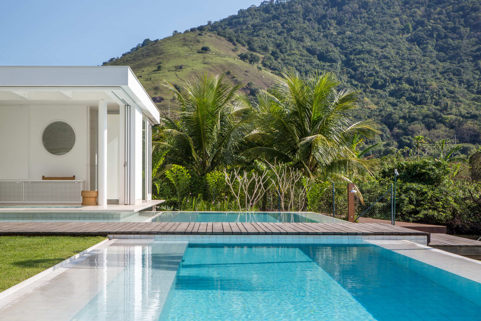 Swimming pool with timber deck island surrounded by lawn and palms below a forested hillside