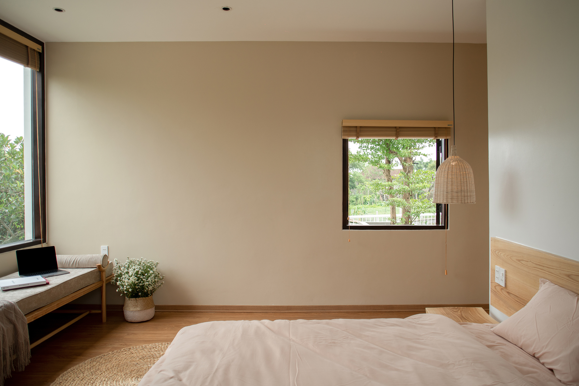 Bedroom with pale walls, timber bed frame, woven pendant light, and window overlooking garden foliage