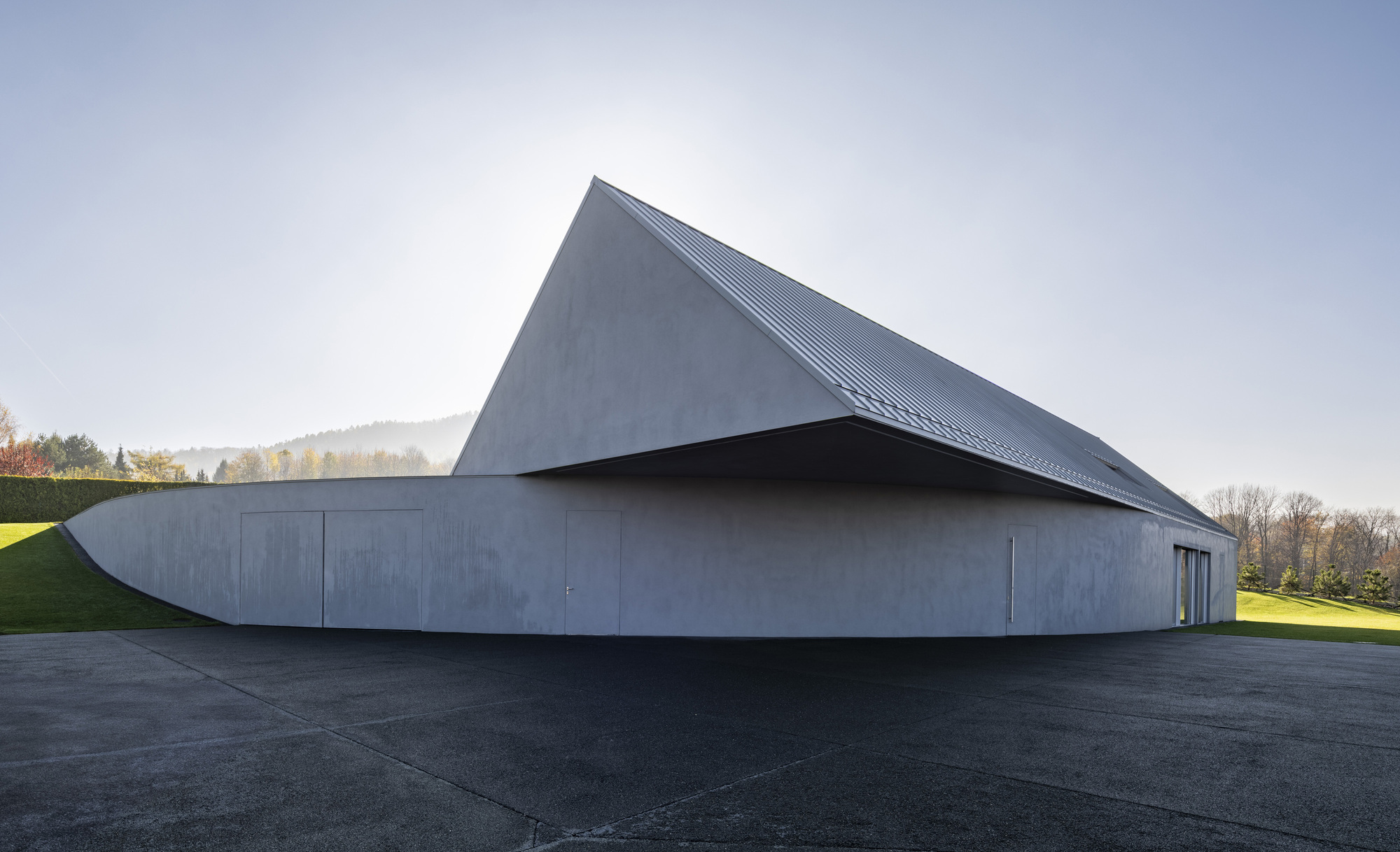 Angular zinc roof volume cantilevering over curved concrete base with flush doors facing the lawn
