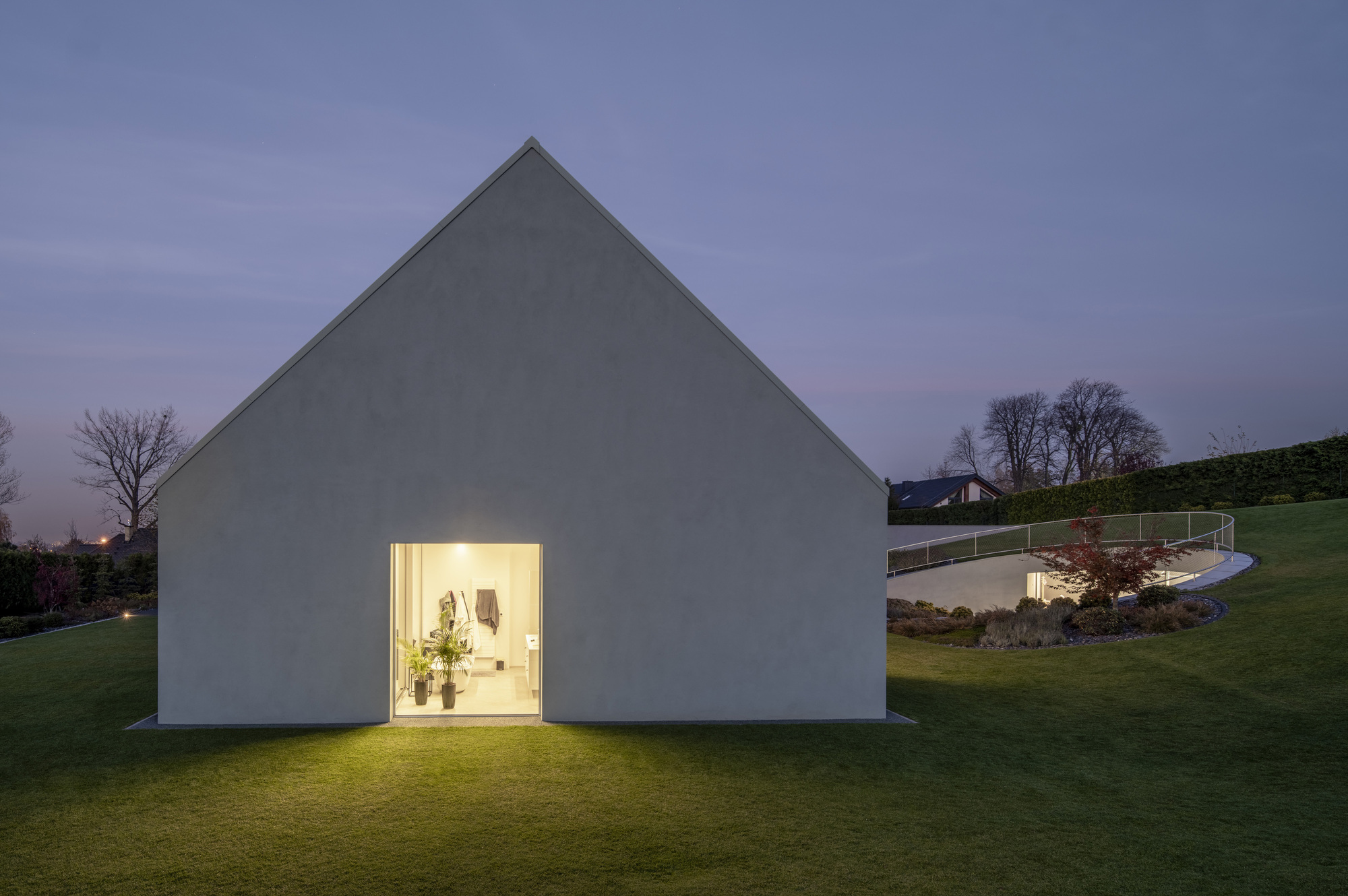 Gabled white volume with single illuminated doorway opening onto lawn at dusk