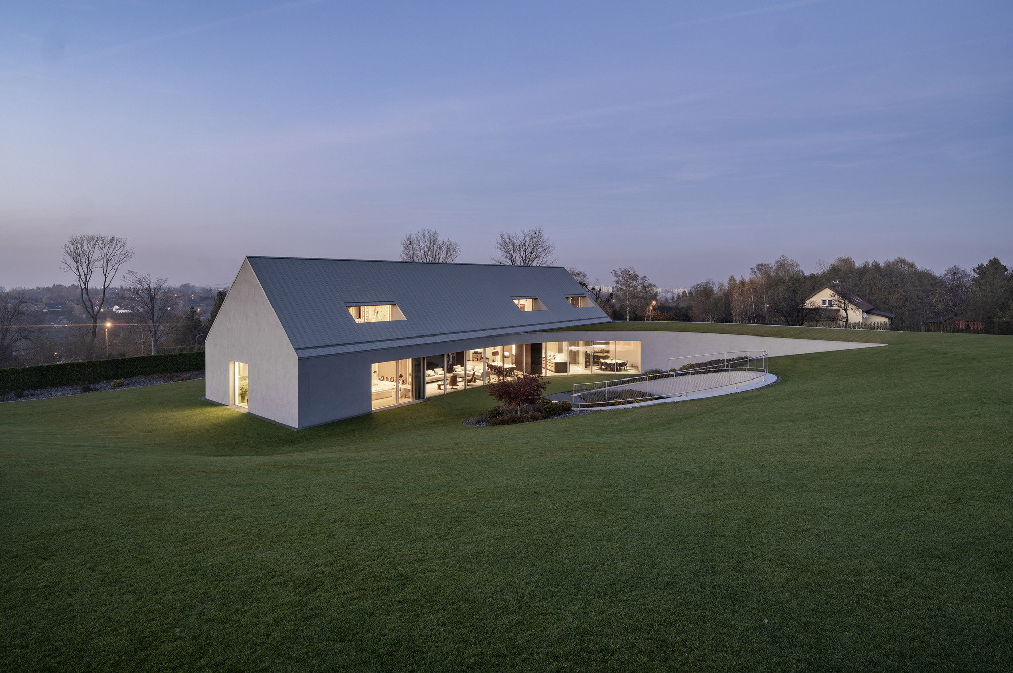 Gabled house with metal roof and glazed lower level set into sloping lawn at dusk