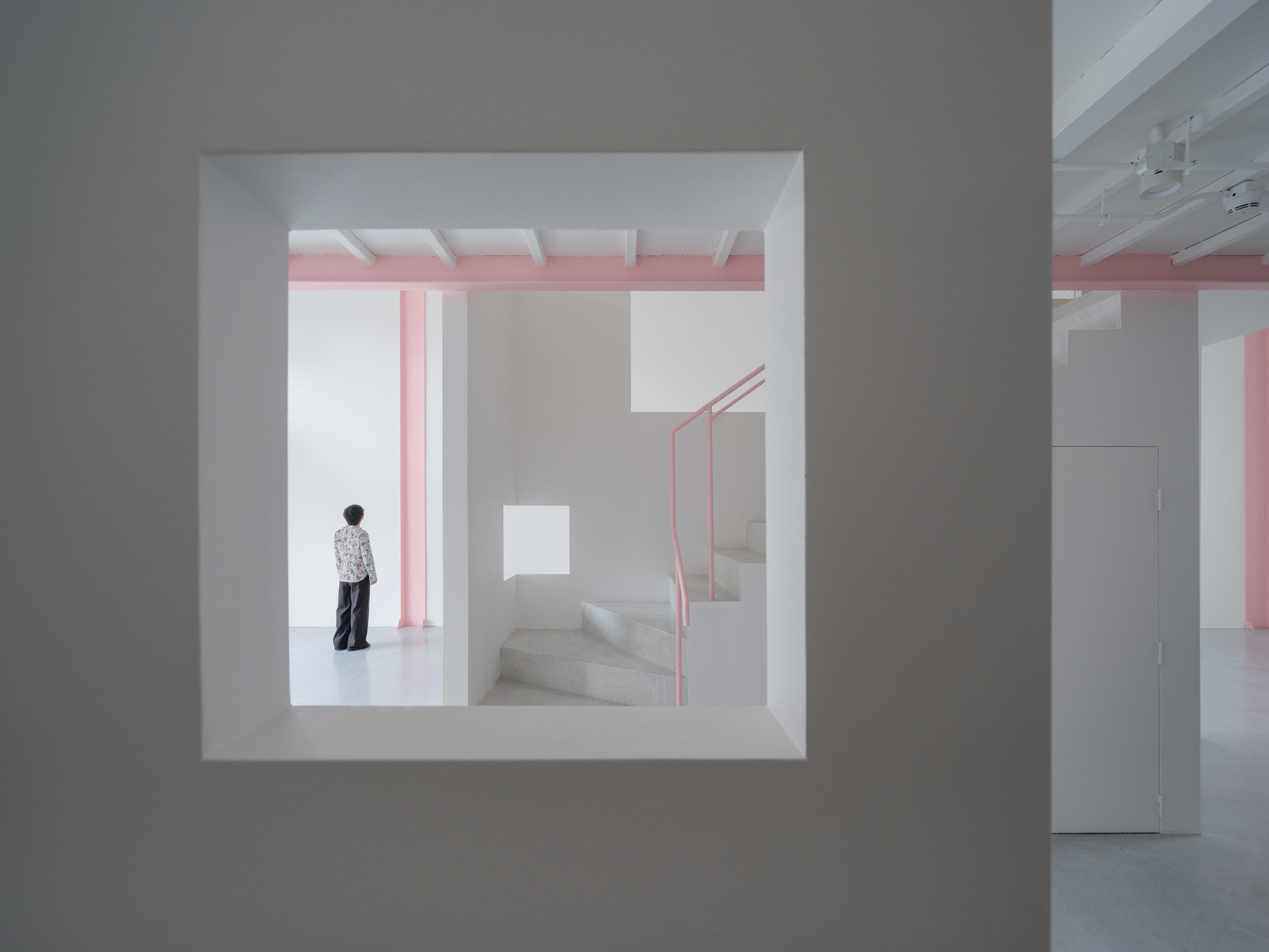 View through a square wall opening showing a person standing near pink steel stairs