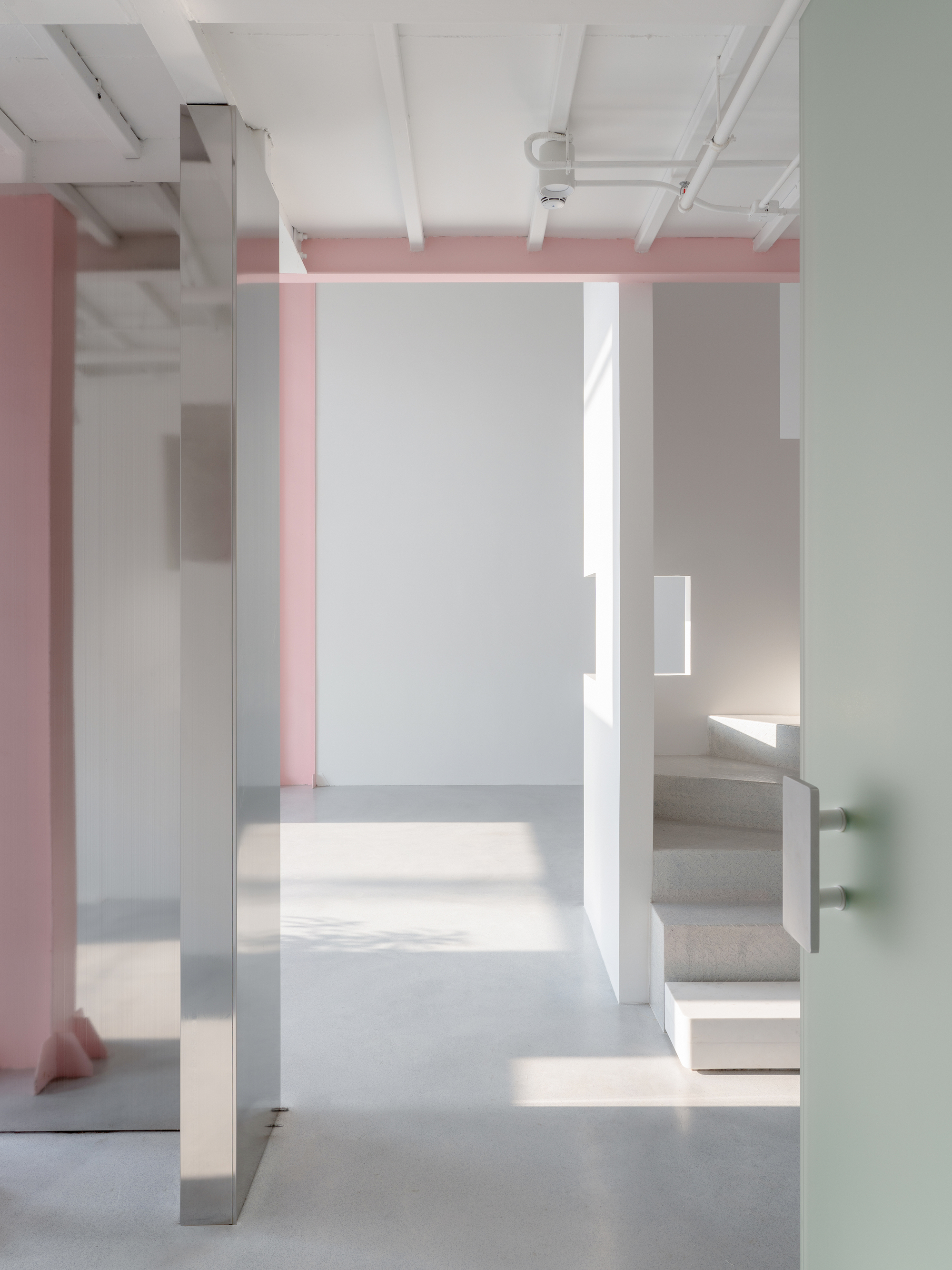Interior view through white partitions with pink beams overhead and sunlight casting geometric shadows on polished concrete floors