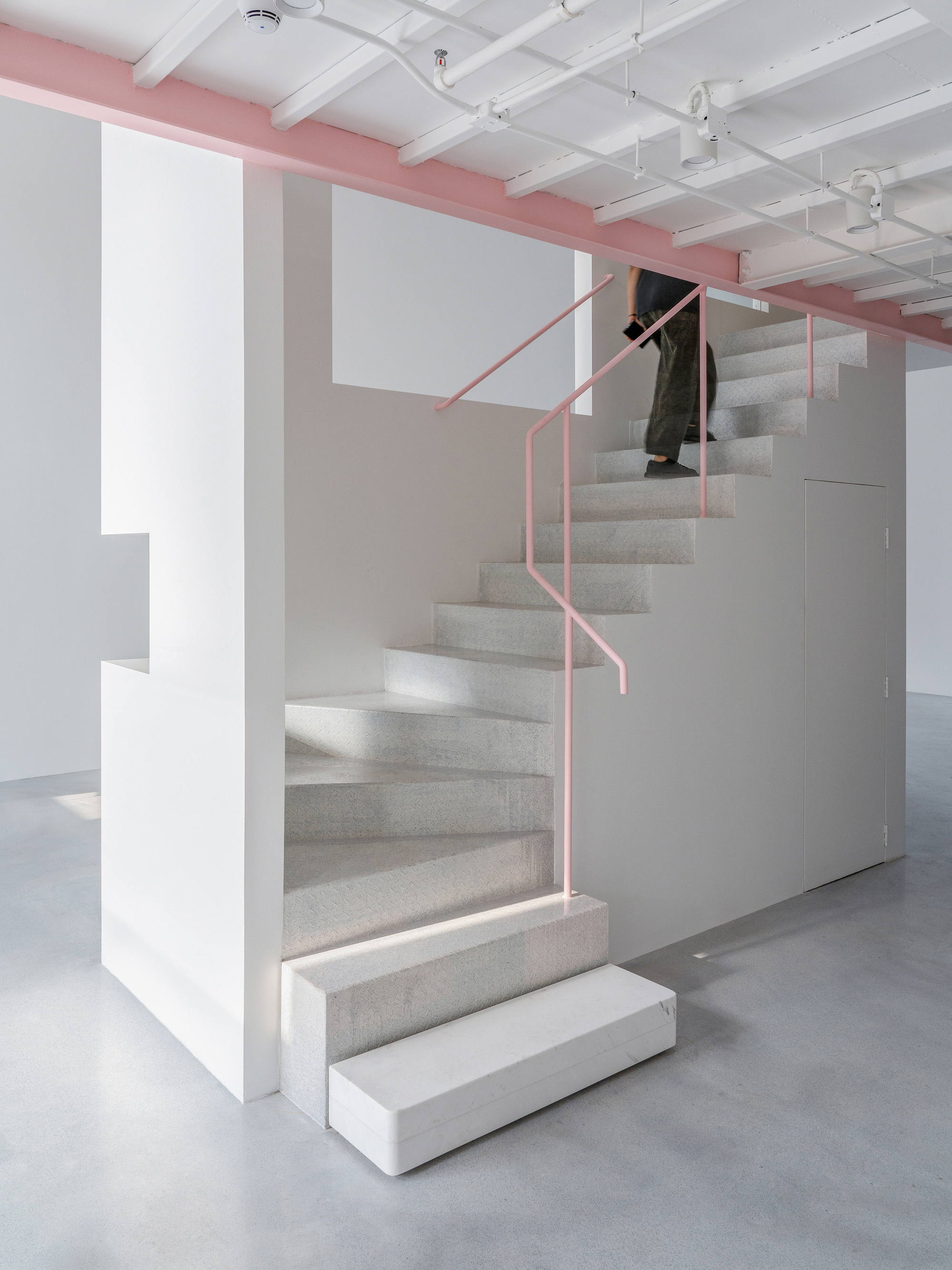 Carpeted staircase with pink tubular steel handrail ascending beneath exposed white coffered ceiling with pink structural beam