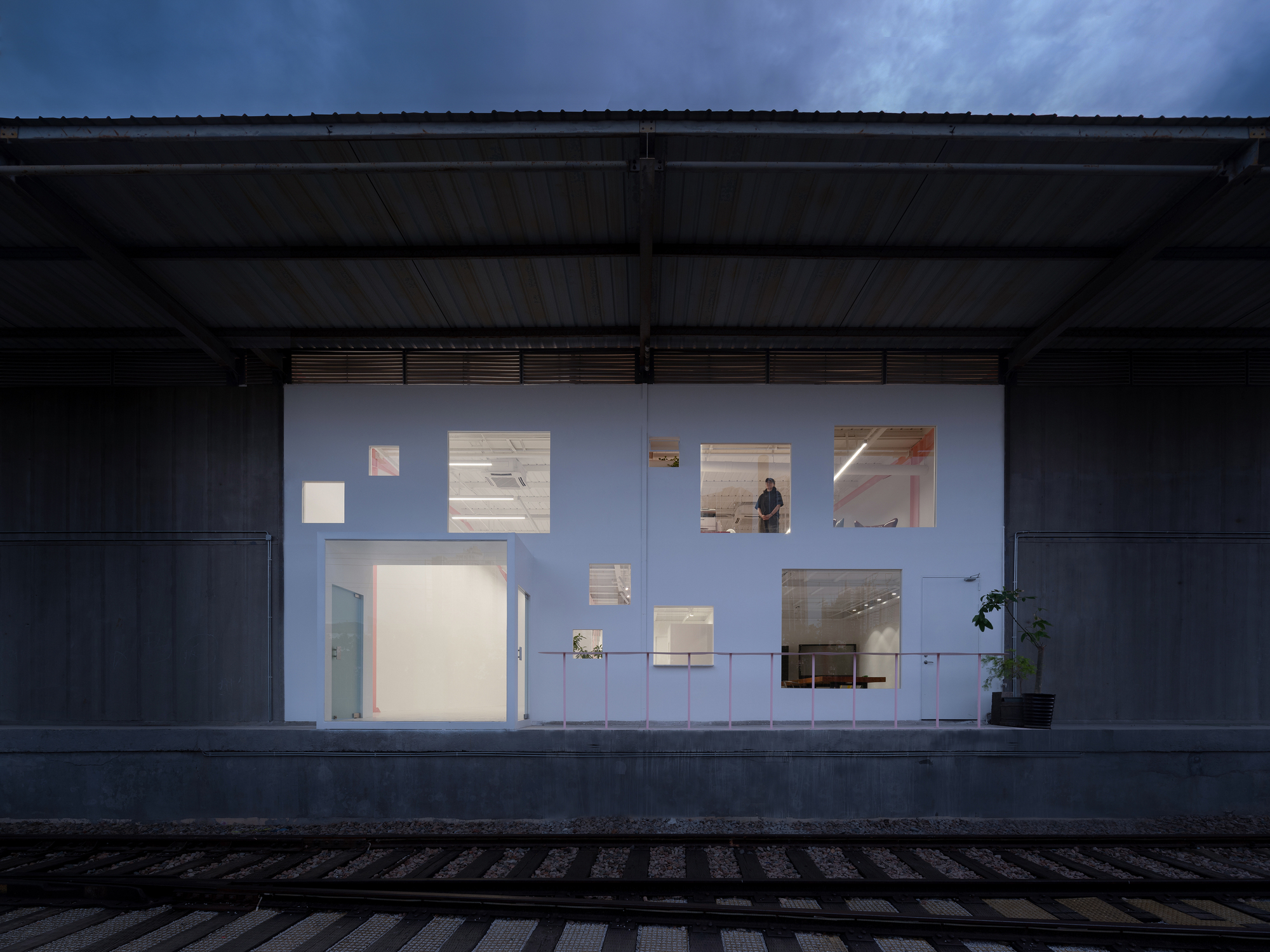 Illuminated white facade wall beneath a metal shed canopy at dusk with a figure visible inside