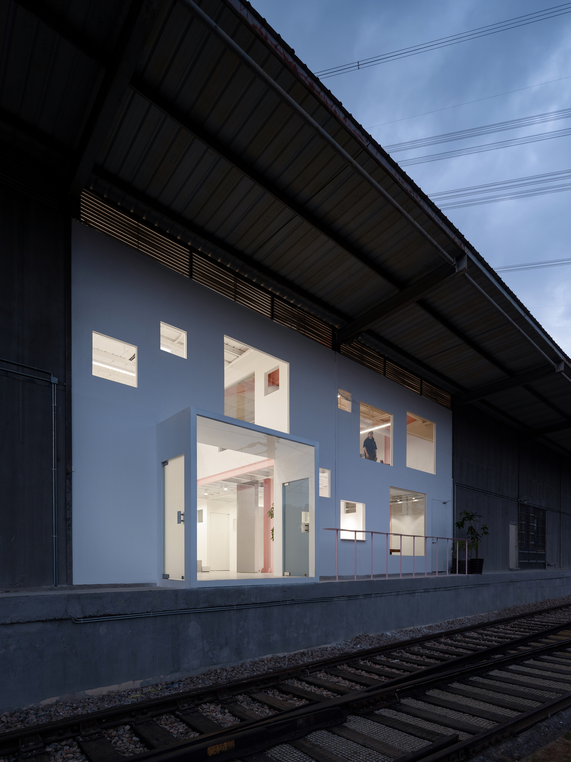 White facade wall with scattered window openings under a corrugated metal canopy and railway tracks in foreground