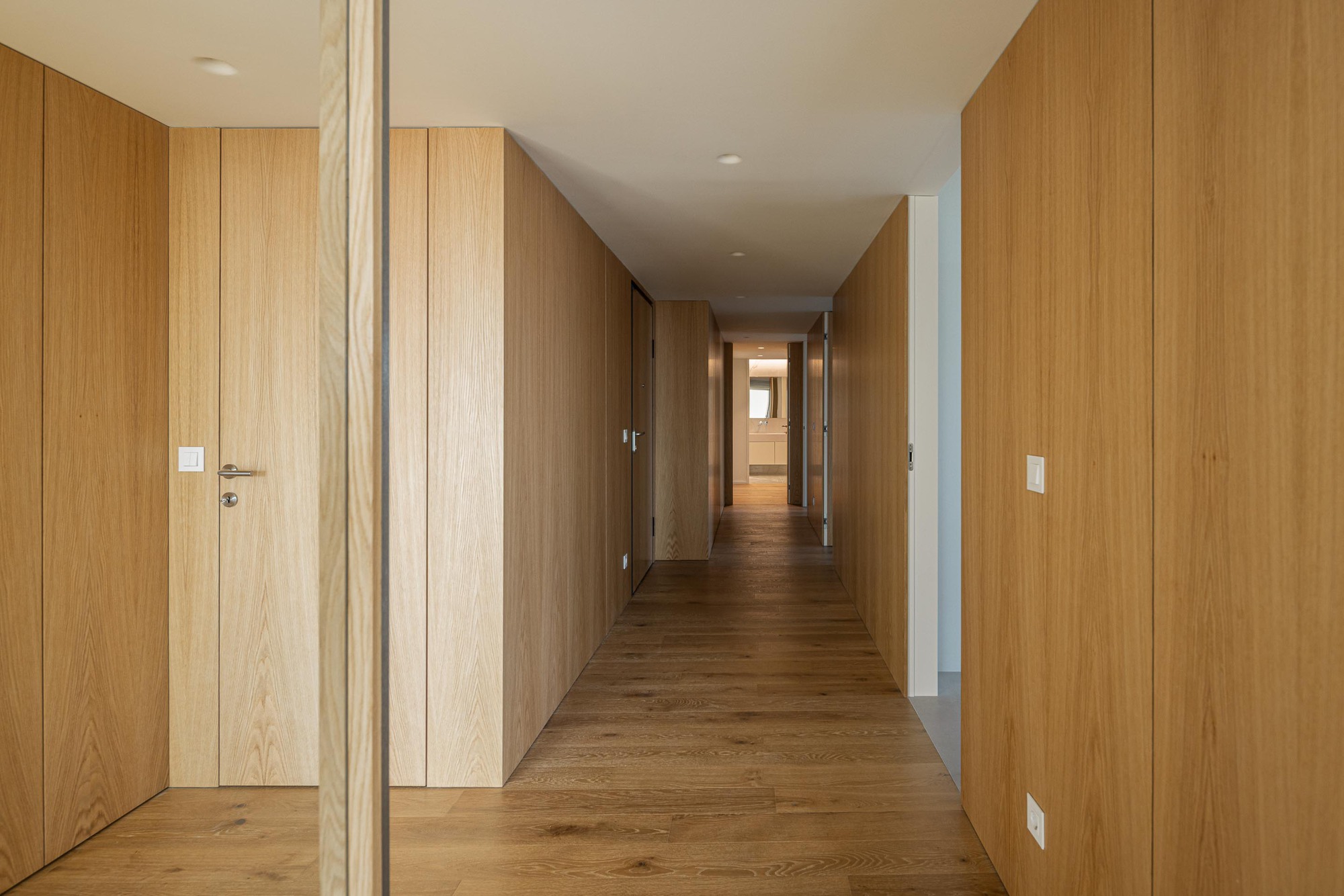 Long corridor lined with continuous timber wall paneling and flush doors extending toward a sunlit end