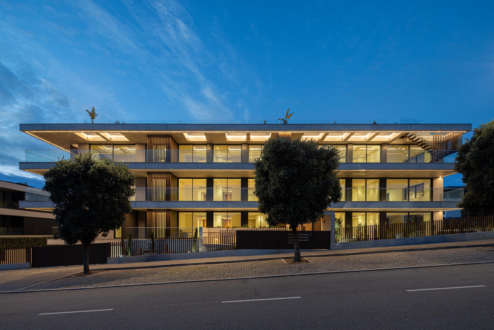 Street view of the illuminated facade with horizontal timber screens and glass balconies at dusk