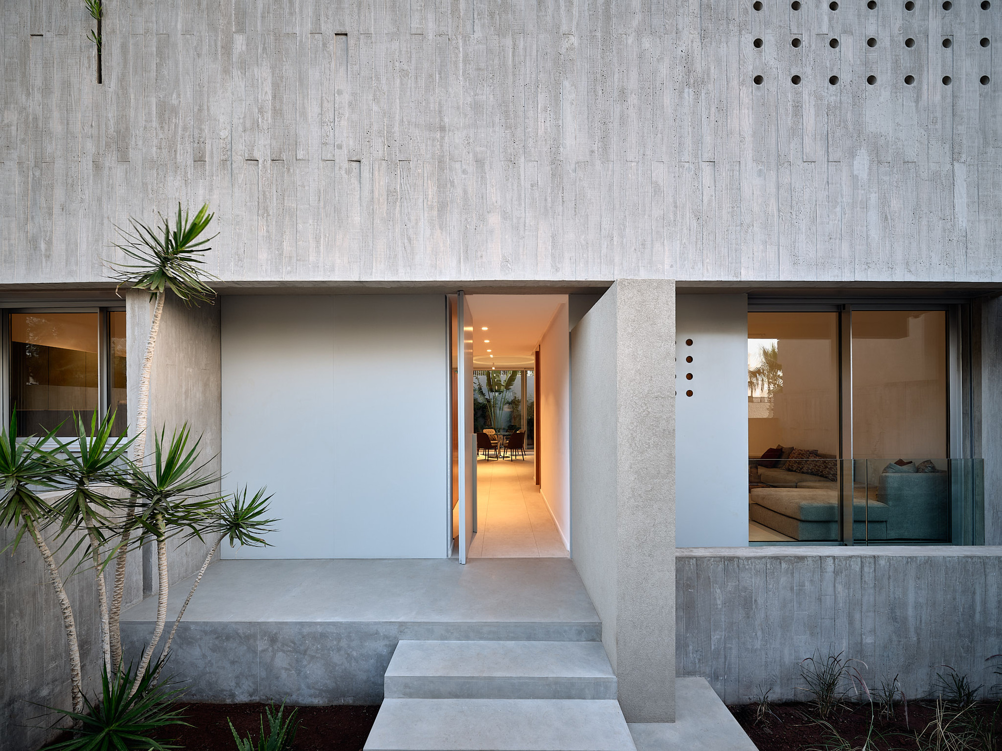 Concrete facade with recessed entry and glass sliding doors flanked by planted beds with yucca at dusk