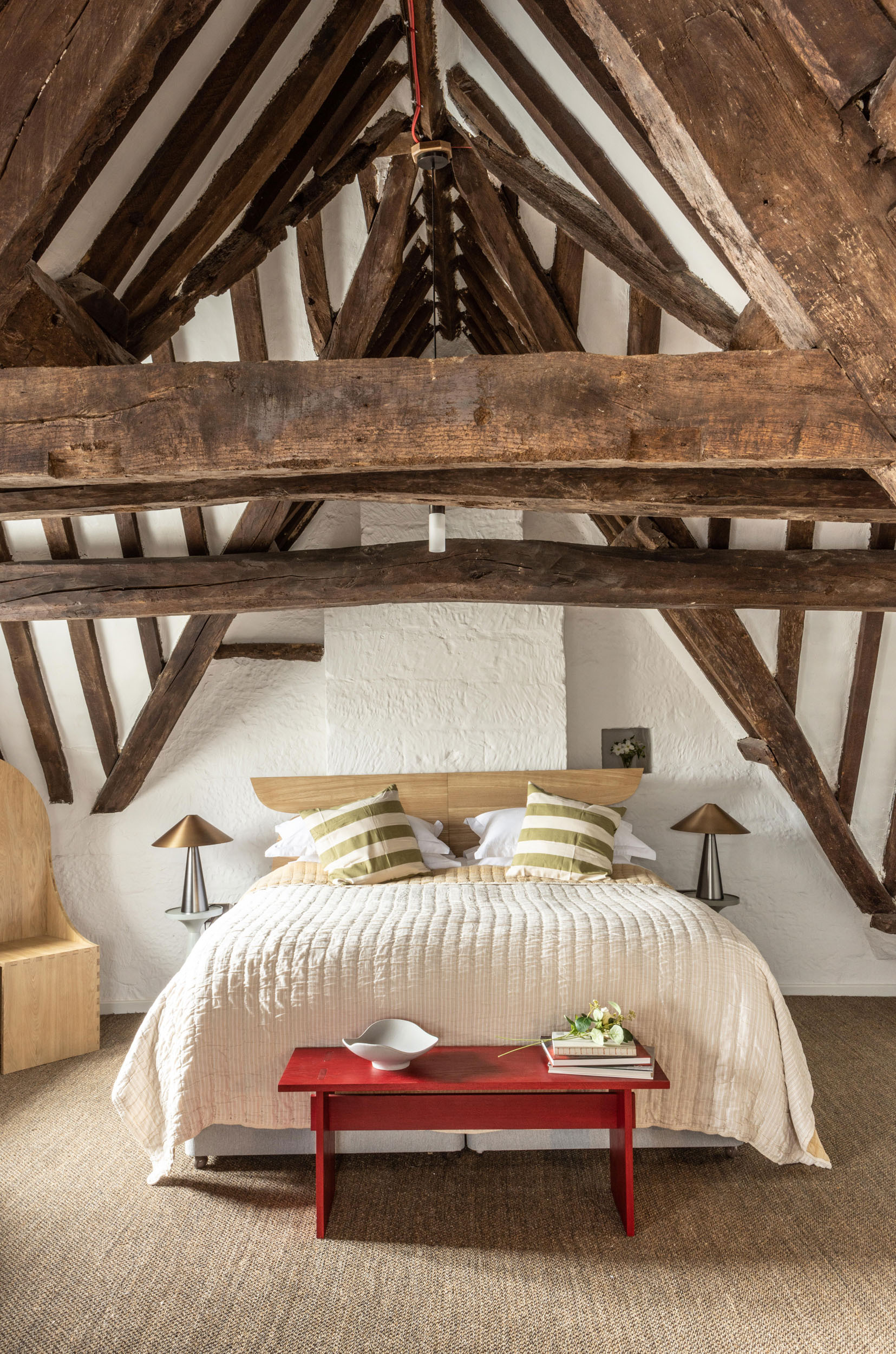 Bedroom beneath exposed timber roof trusses with a red bench at the foot of the bed