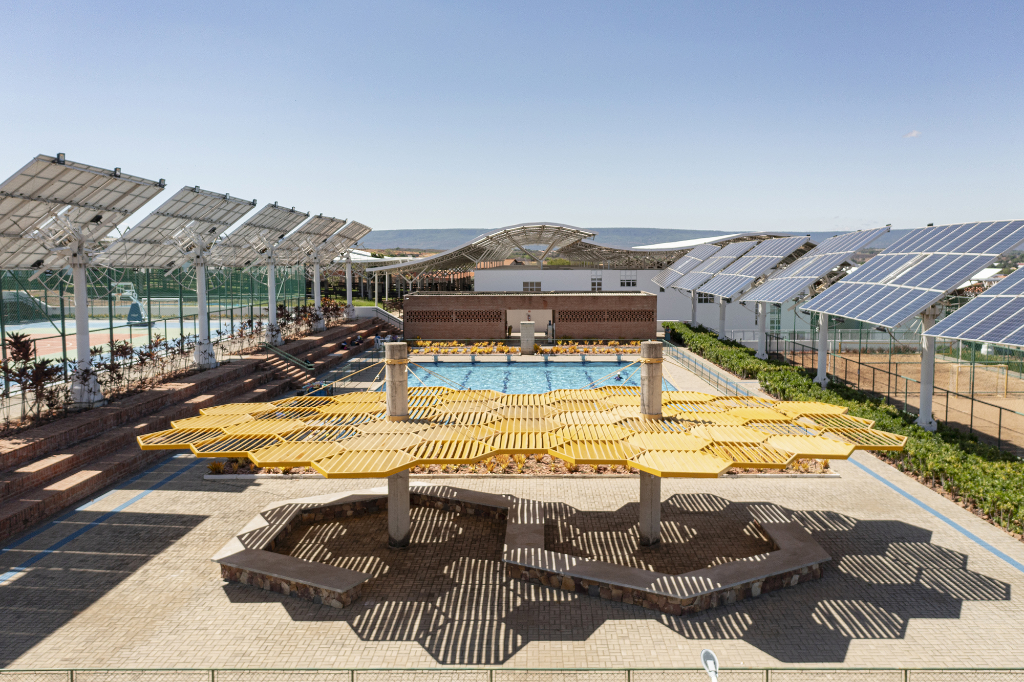 Yellow timber canopy structure casting geometric shadows over amphitheater seating and pool area