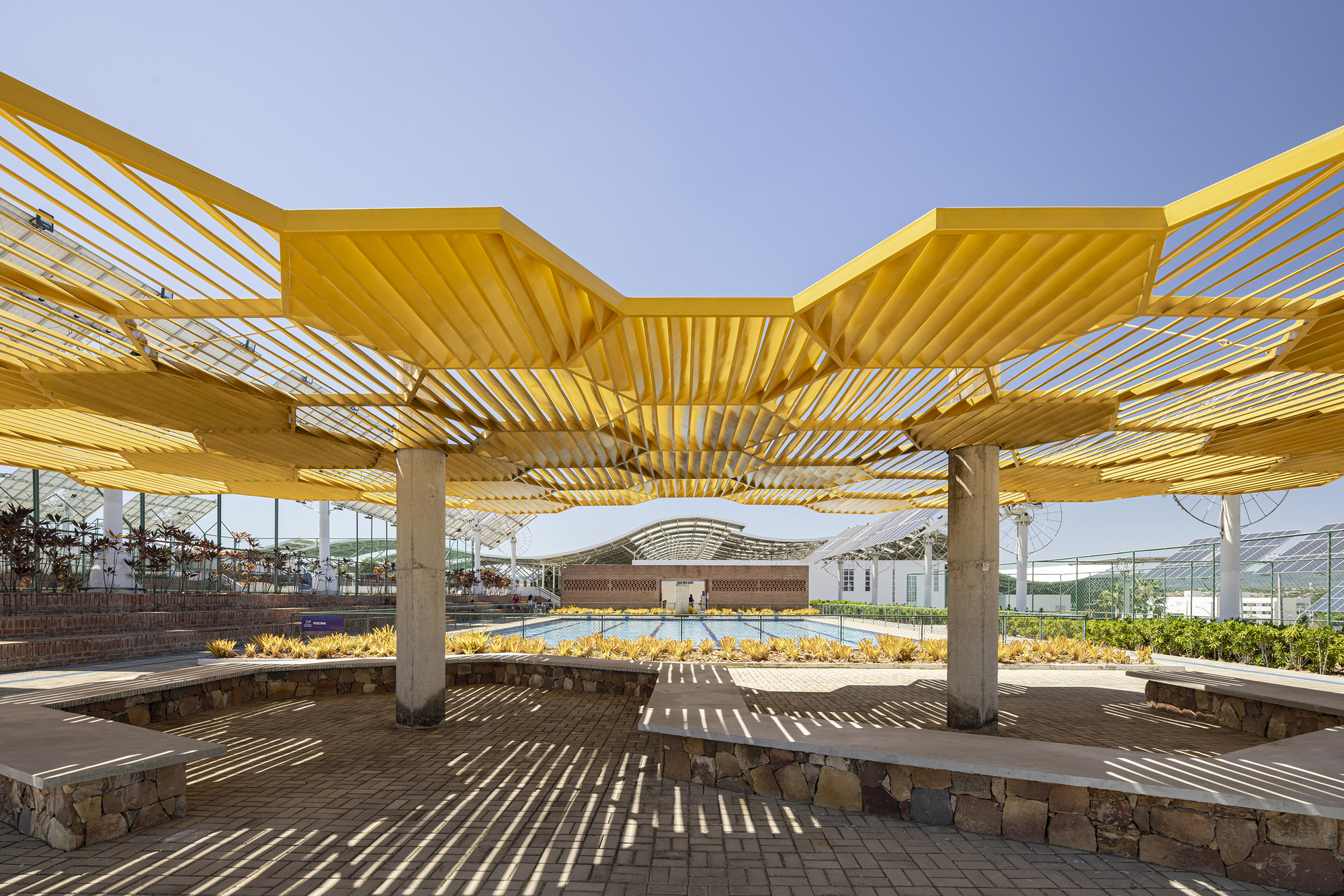 Seating area beneath the yellow slatted pergola with concrete columns and stone bench walls