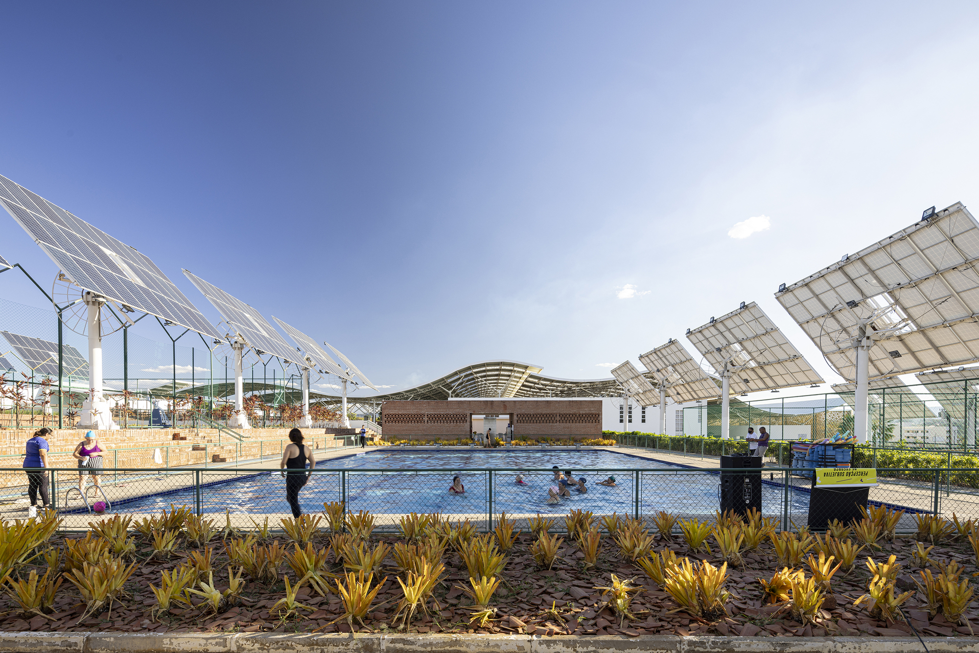 Outdoor swimming pool with fenced perimeter and overhead solar panel array under clear blue sky