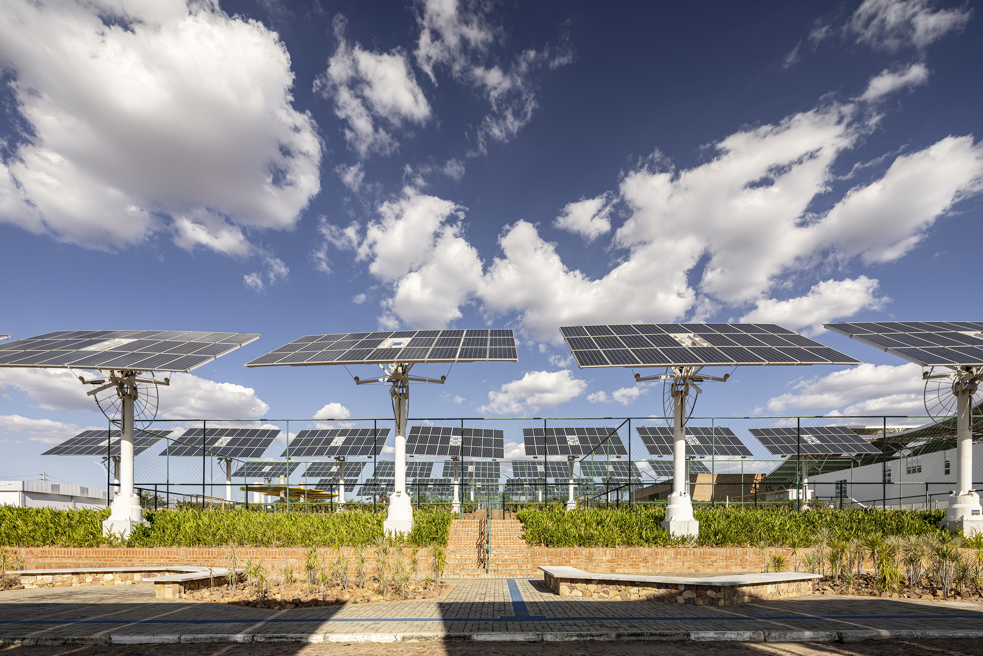 Row of tilted solar panels on white columns with pathway and scattered clouds overhead