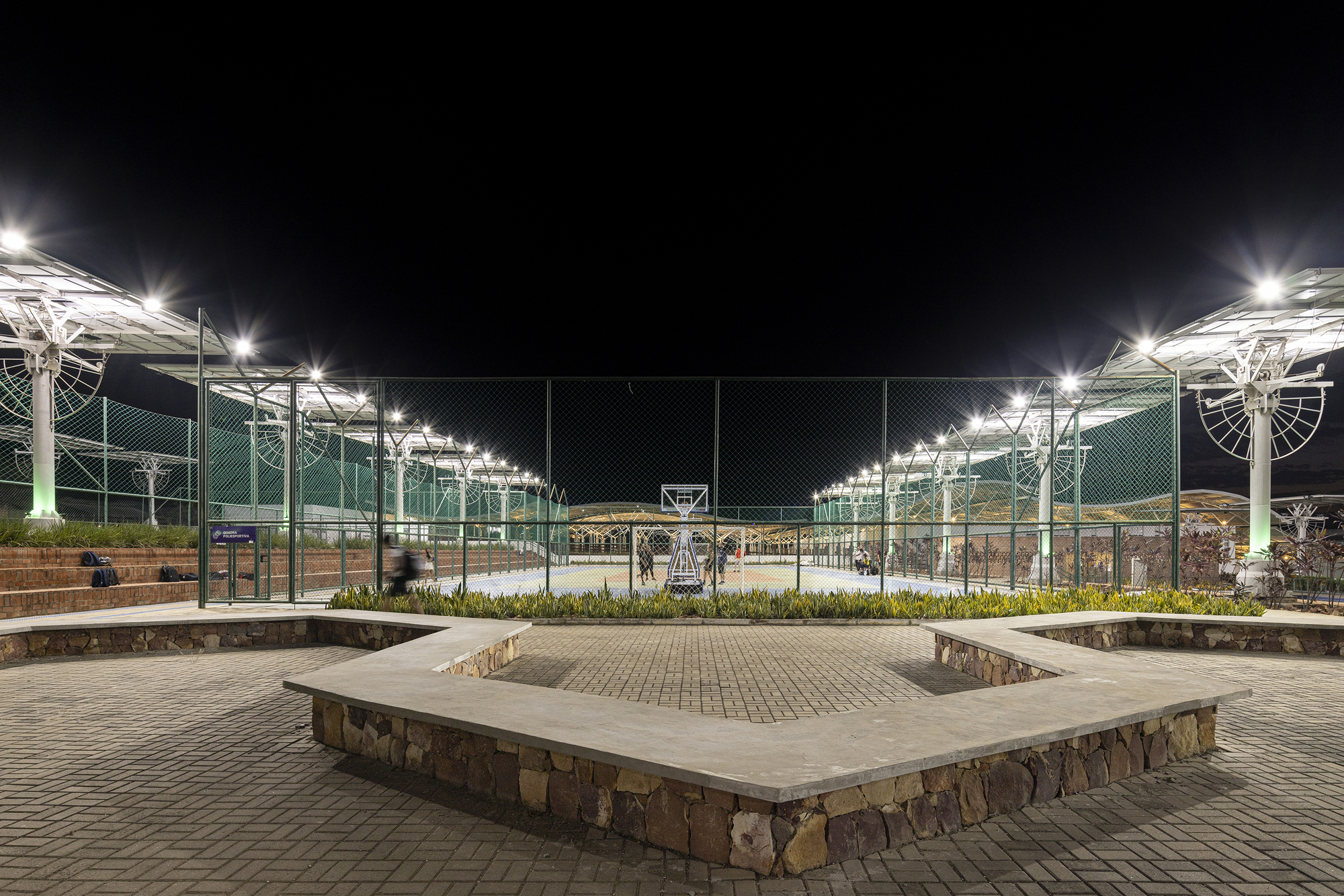 Stone and concrete seating platform with metal fencing and lit solar canopies beyond at night