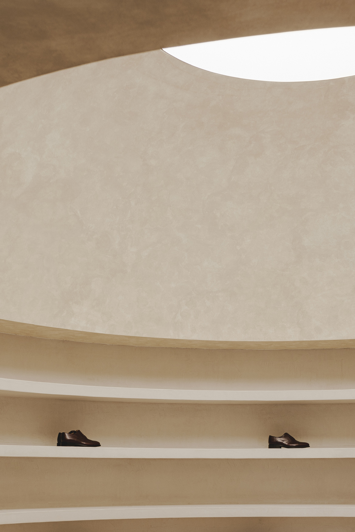 Curved plaster wall with circular skylight above continuous timber shelving holding two shoes