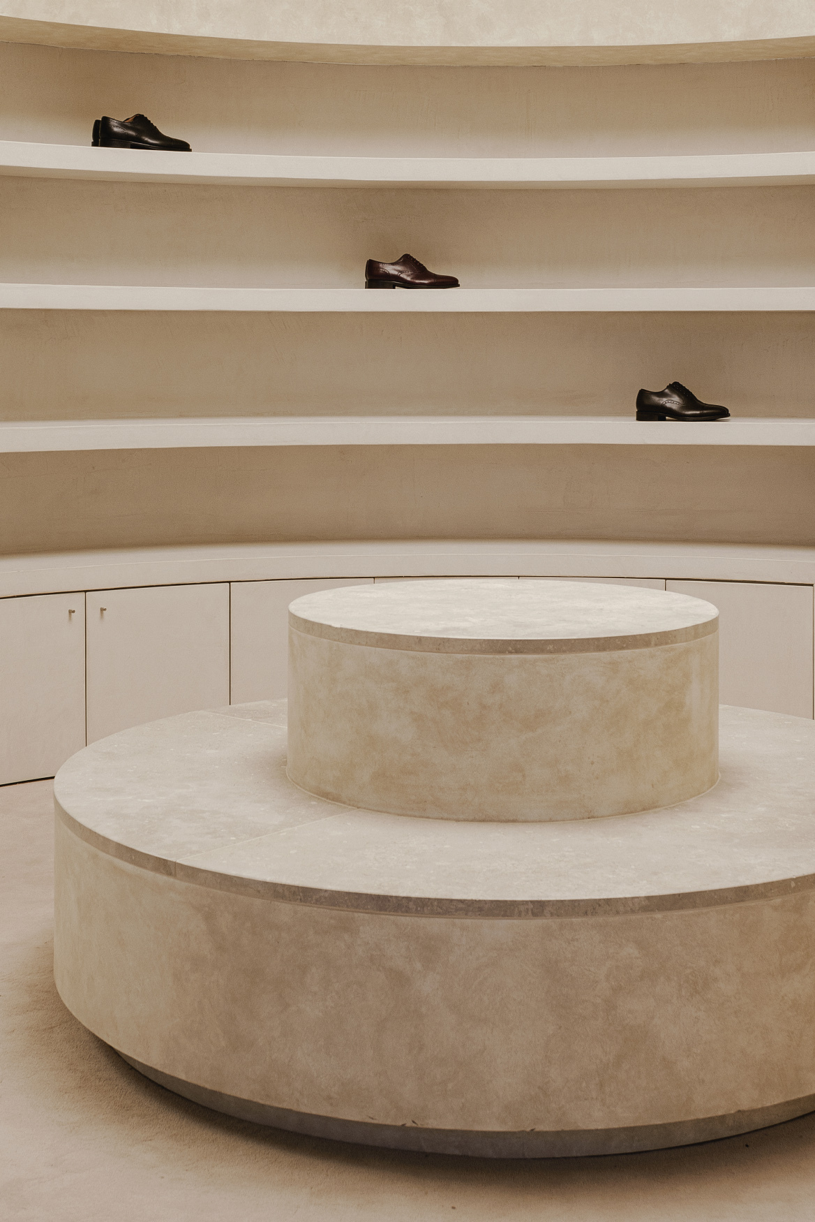 Tiered circular plinth in limestone below curved wall with shelving displaying three shoes