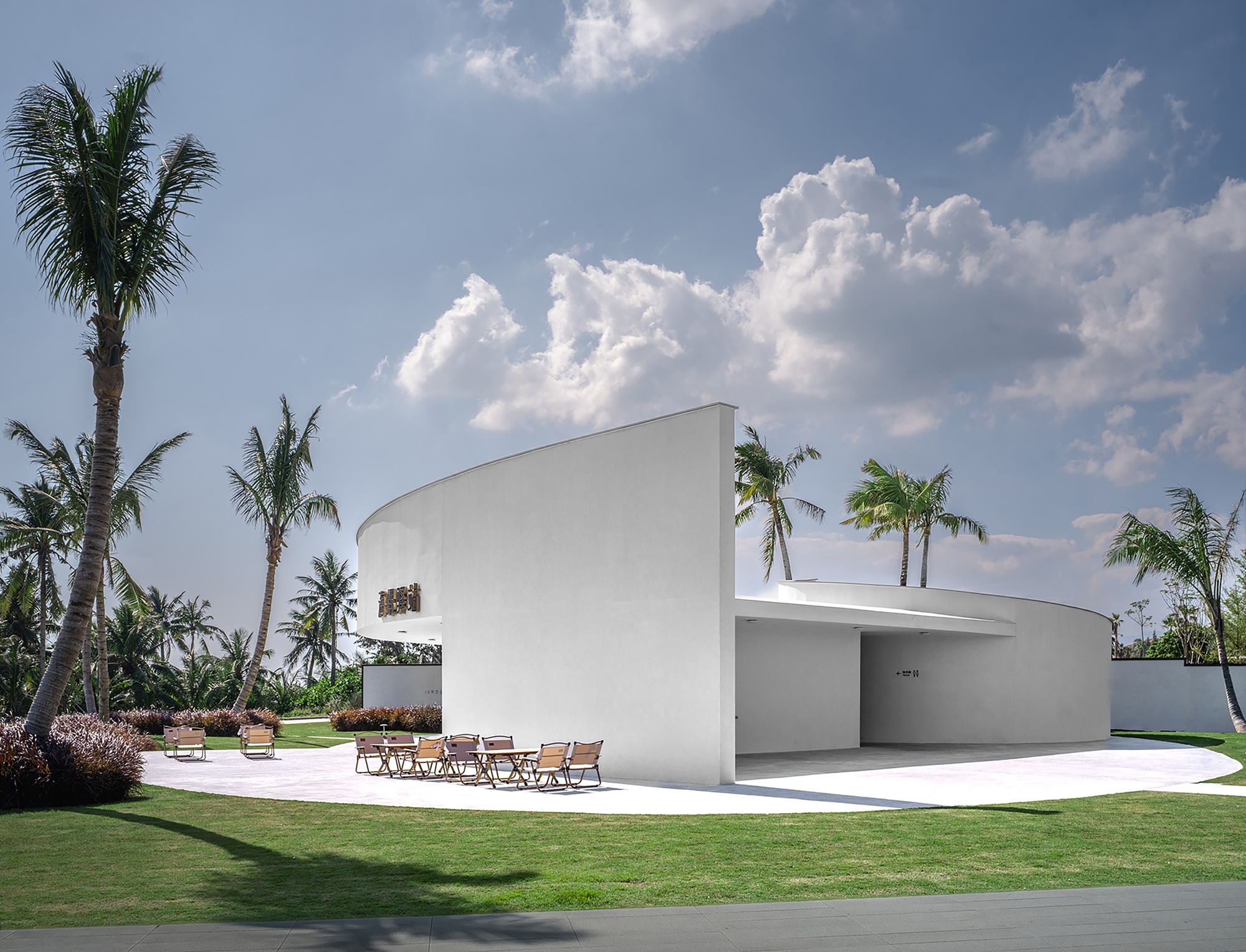 White curved pavilion with cantilevered roof and outdoor terrace surrounded by palm trees and manicured lawn