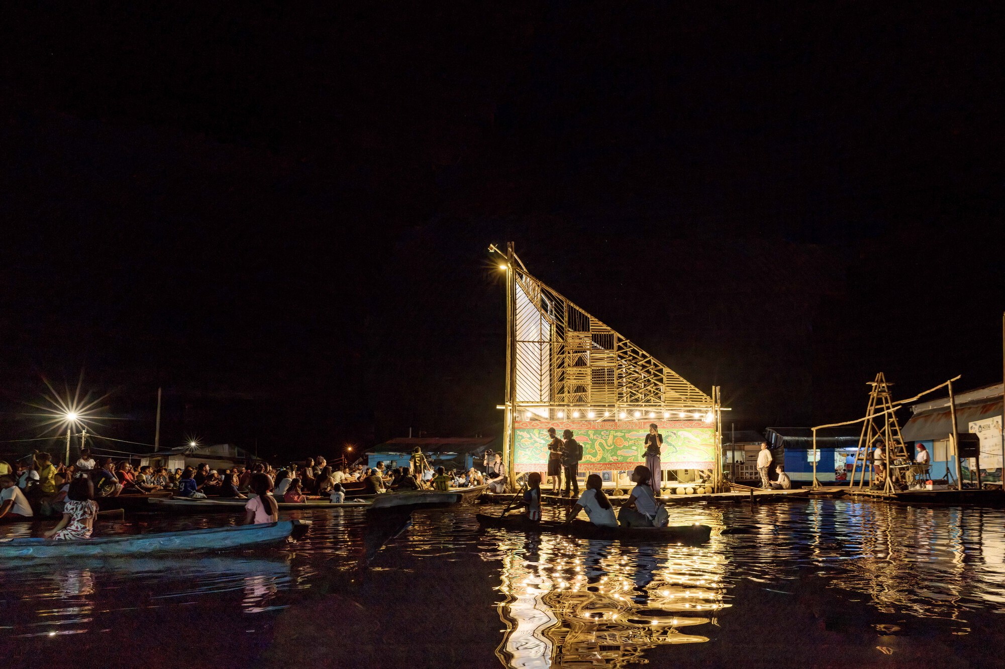 Illuminated bamboo lattice screen and projection wall reflected in dark water with boats nearby at night