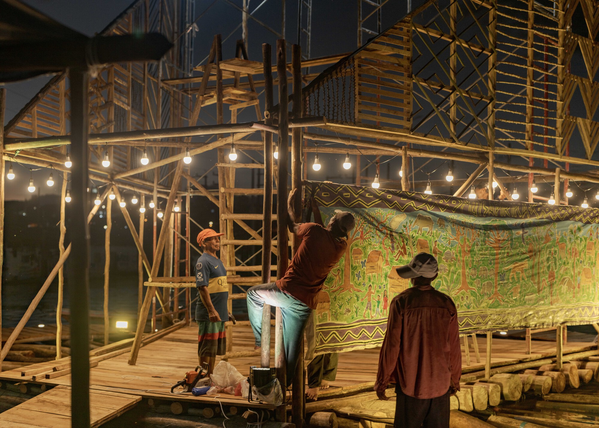 Timber scaffold structure with hanging bulb lights and a painted textile backdrop at dusk