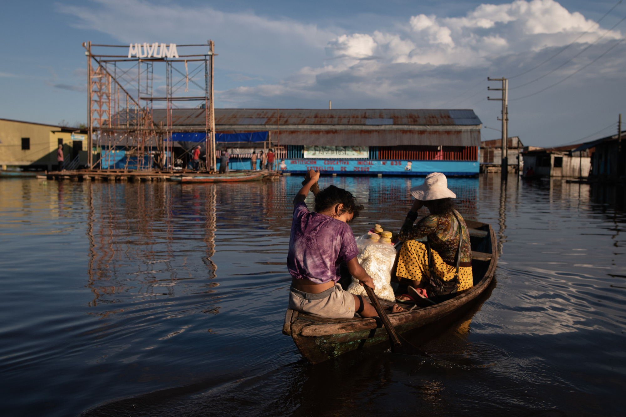 Floating blue school structure viewed from a wooden boat approaching across flooded waters in late afternoon