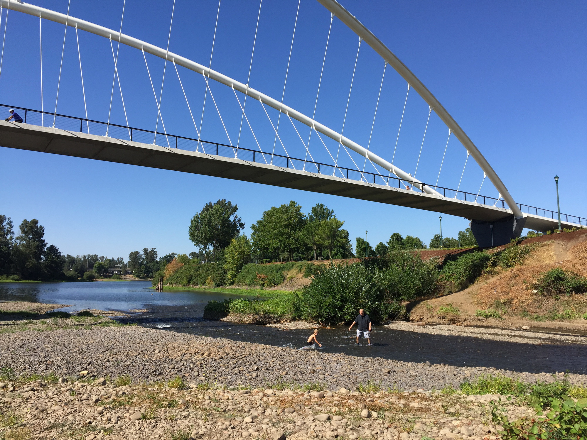 Bridge spanning river with person walking on gravel path along the water's edge below