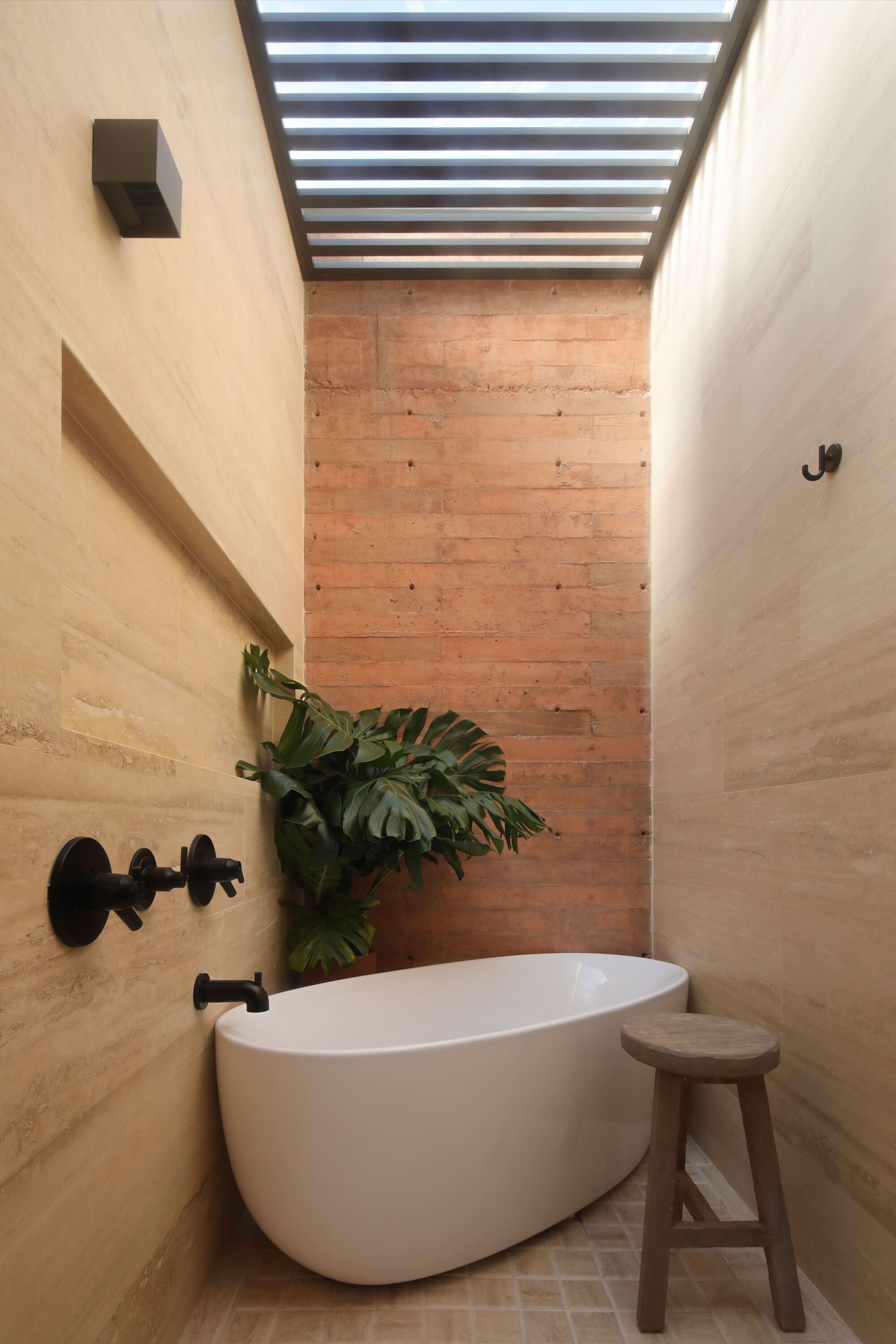 Bathroom with freestanding tub beneath a horizontal slatted skylight and rammed earth wall