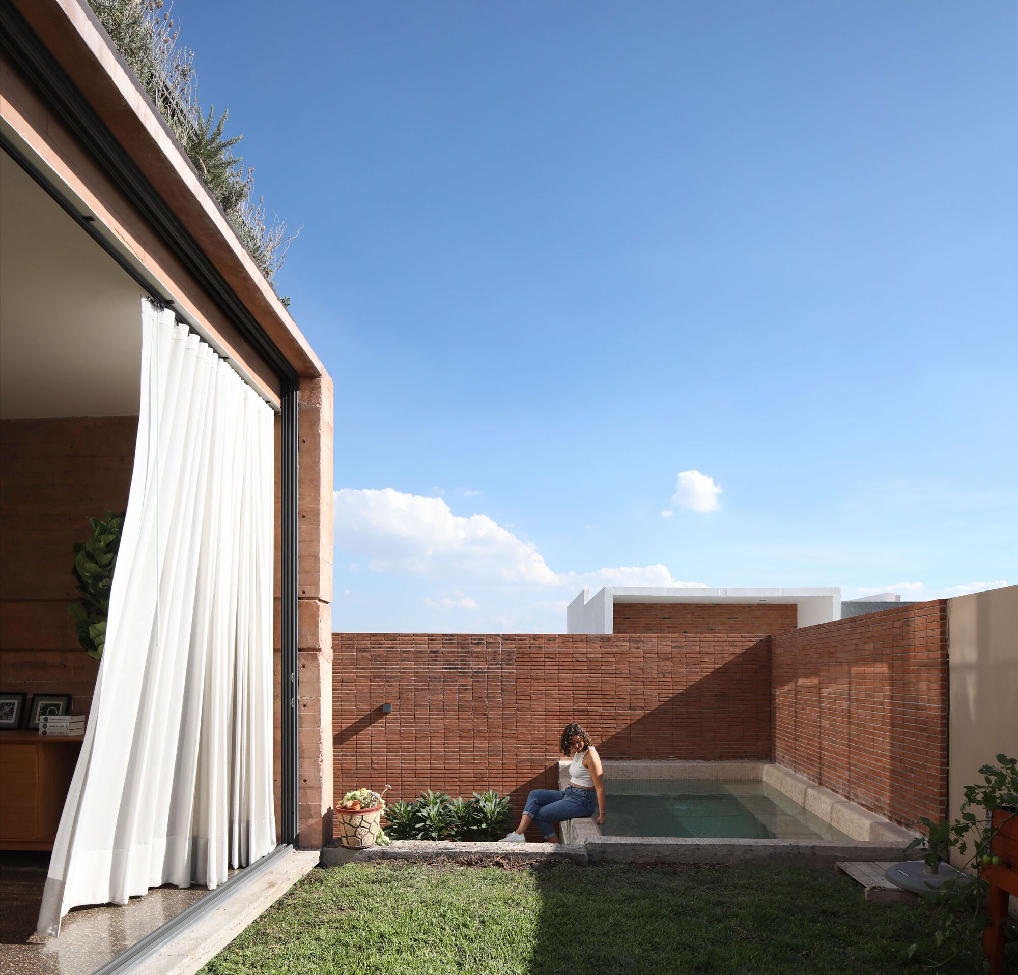 Private courtyard with brick walls, lawn, pool and person seated beside a planted bed under blue sky