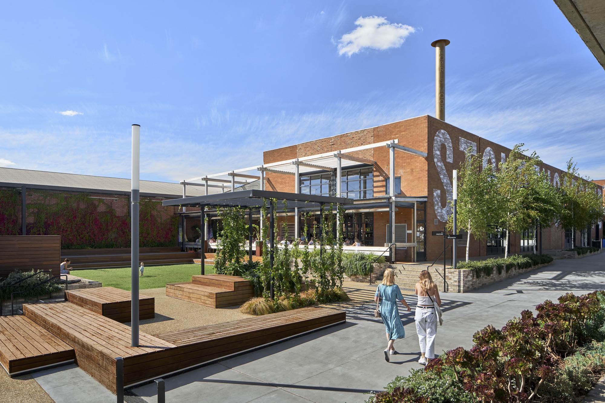 Courtyard with timber-edged planters and steel-framed canopy structures beneath a blue sky with scattered clouds