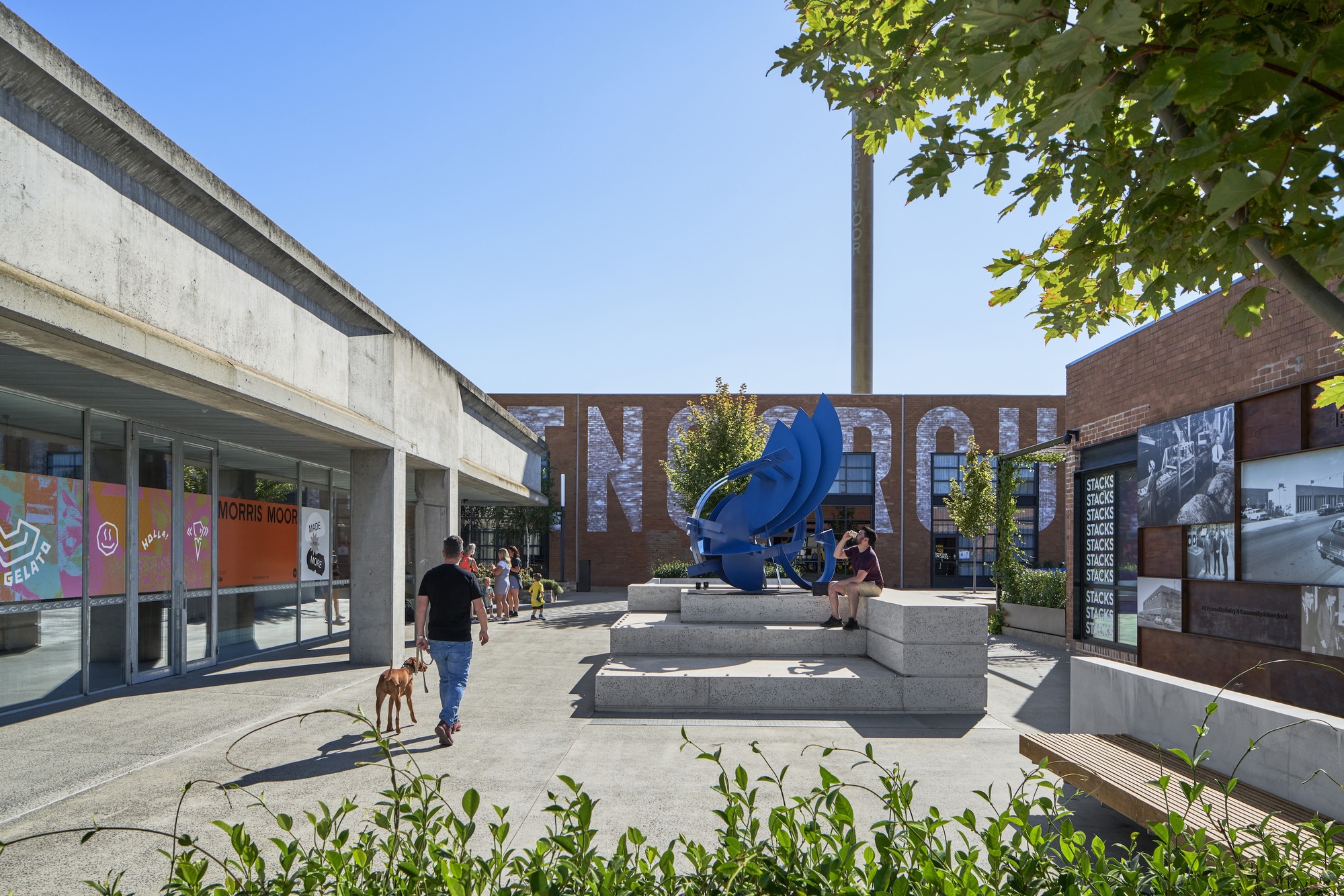 Public plaza with blue sculpture on concrete steps and tree canopy overhead