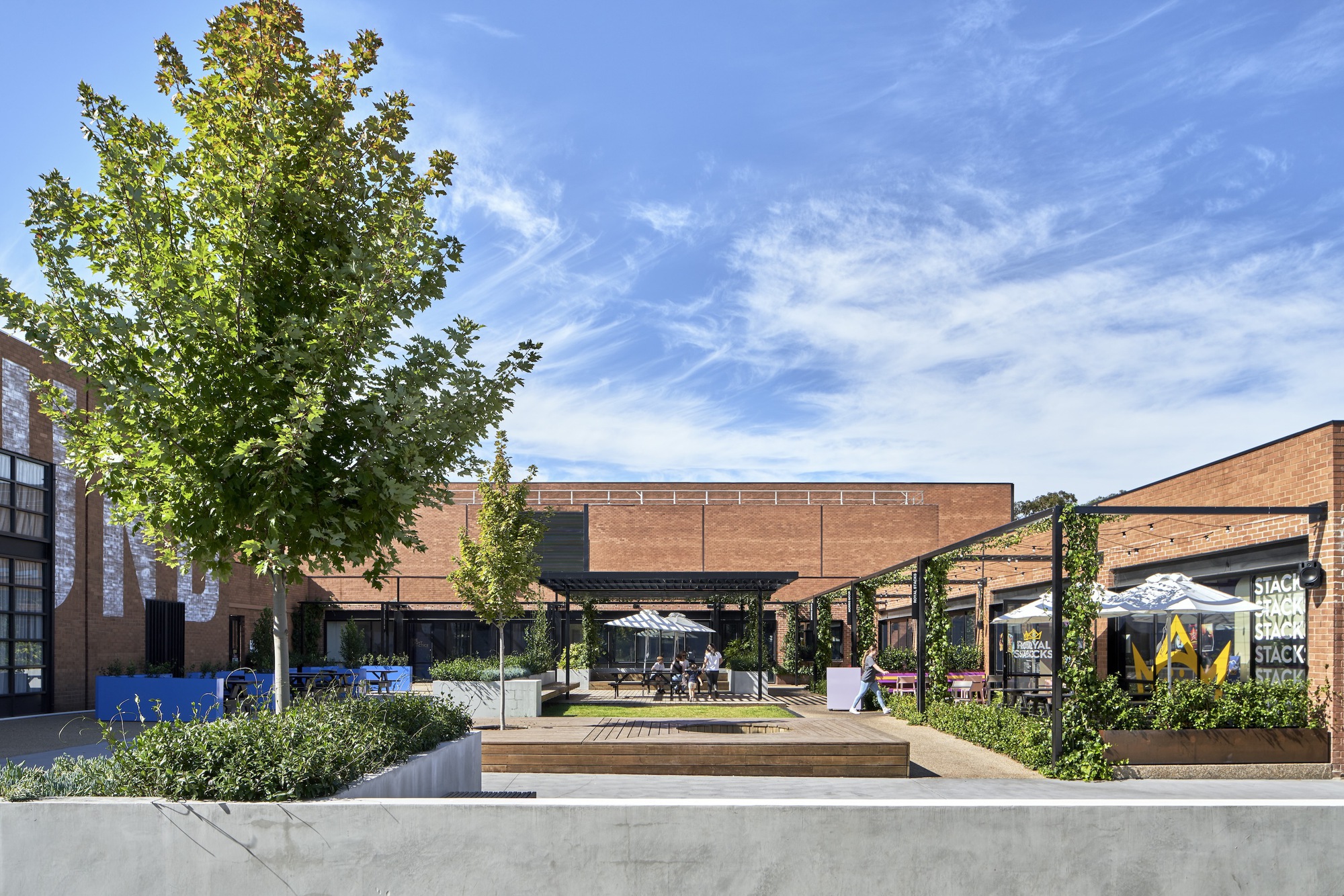 Wide courtyard view showing timber deck, vine-covered steel columns and brick volumes beyond