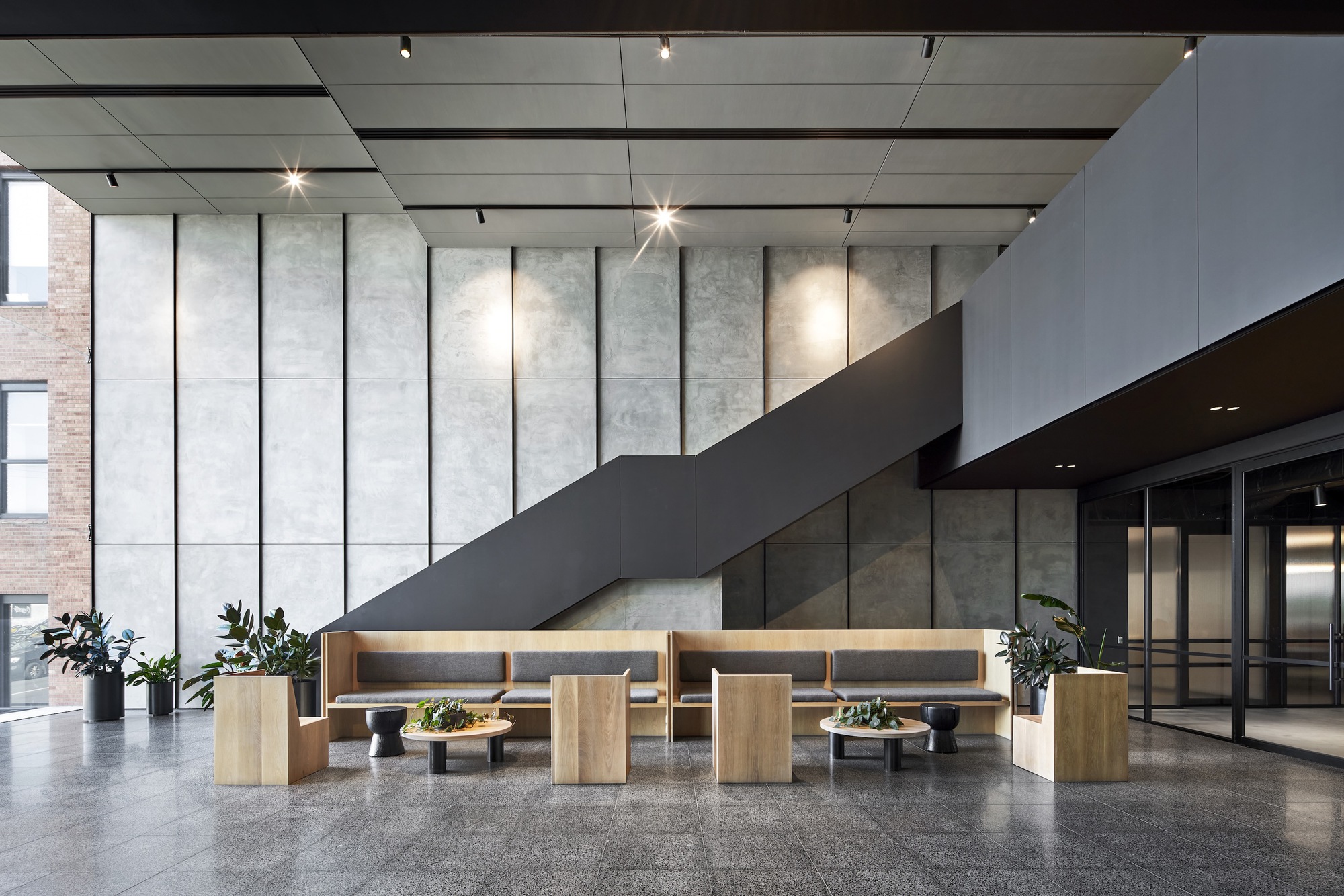 Double-height lobby with gridded concrete wall panels, overhead track lighting, and timber seating banquettes below cantilevered staircase