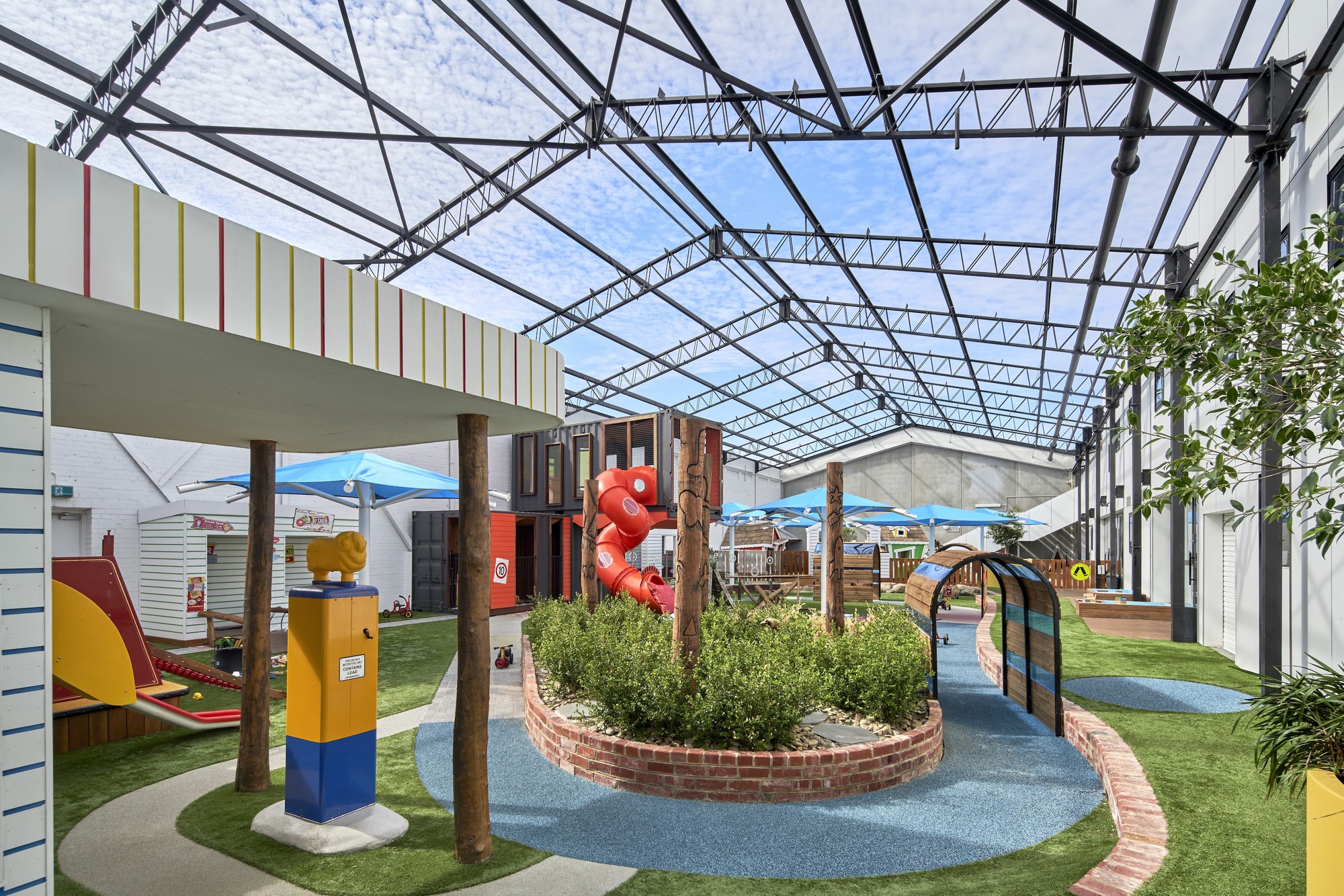 Indoor play area under exposed steel truss roof with planted beds, artificial turf, and timber support columns