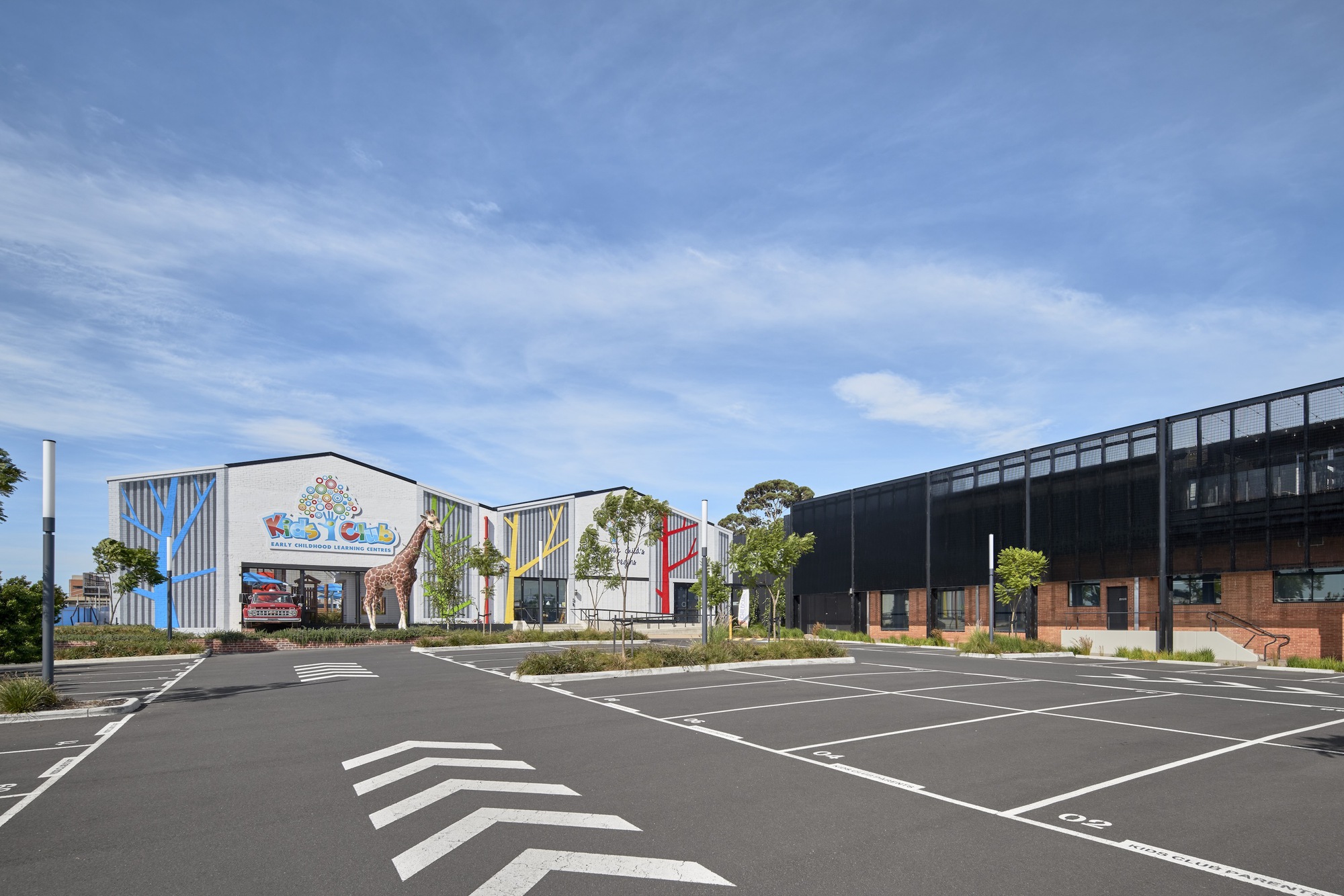 Parking lot view of gabled building cluster with corrugated metal cladding and coloured facade graphics under clear sky