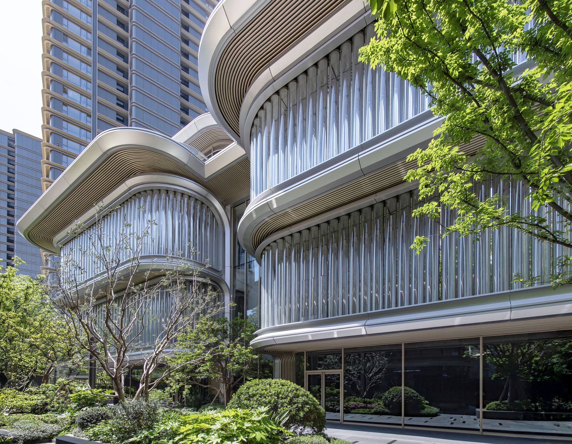 Layered curved balconies with vertical louvers and ribbed soffits framed by trees in sunlight