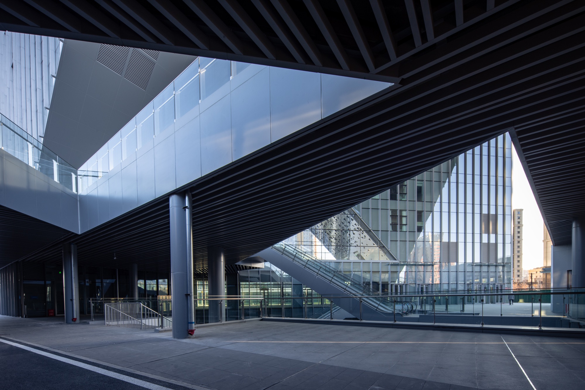 Covered plaza beneath black slatted canopy with escalator and glass facades reflecting twilight sky