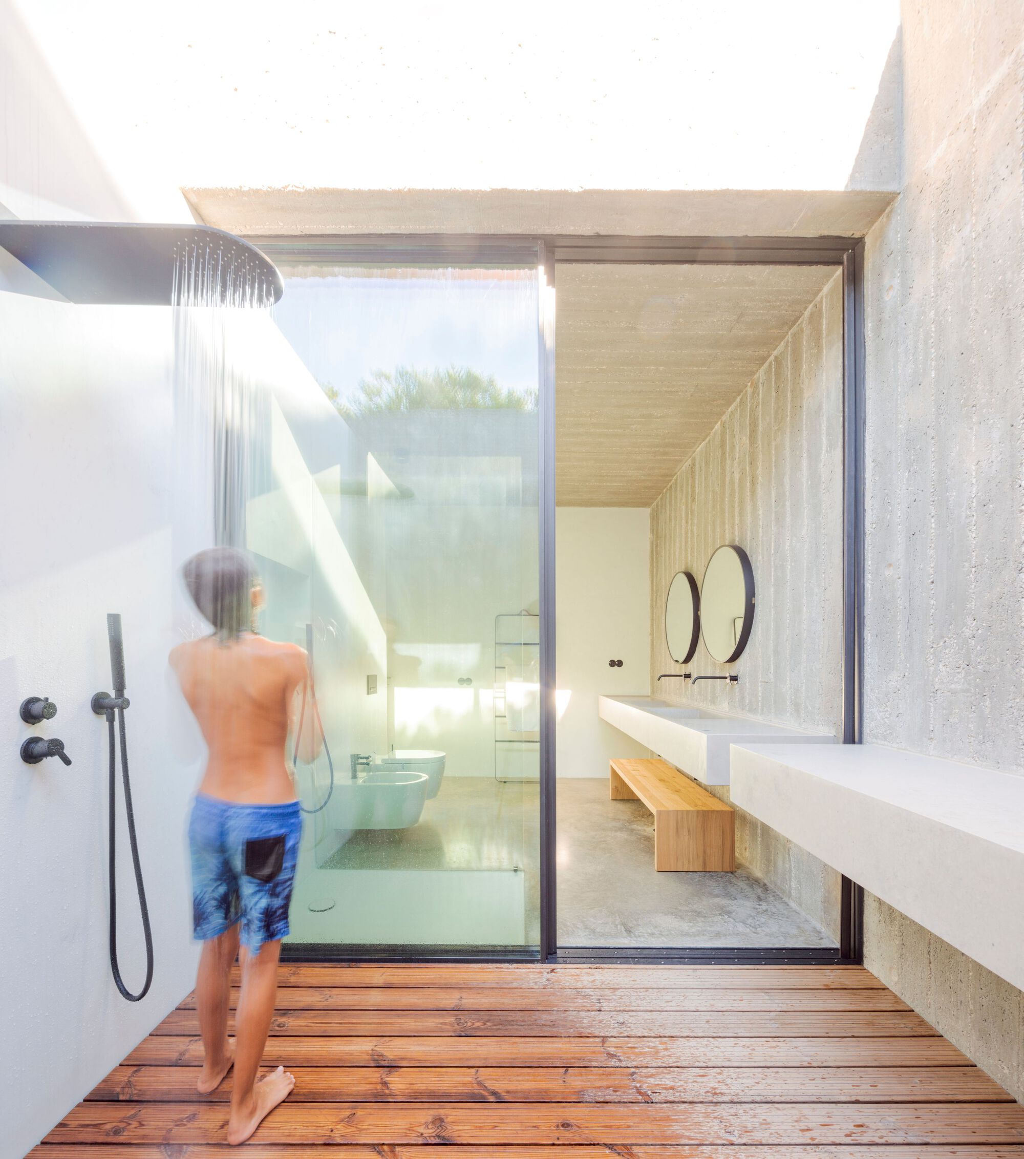 Outdoor shower courtyard with timber decking and figure standing beneath rainfall showerhead in bright sunlight