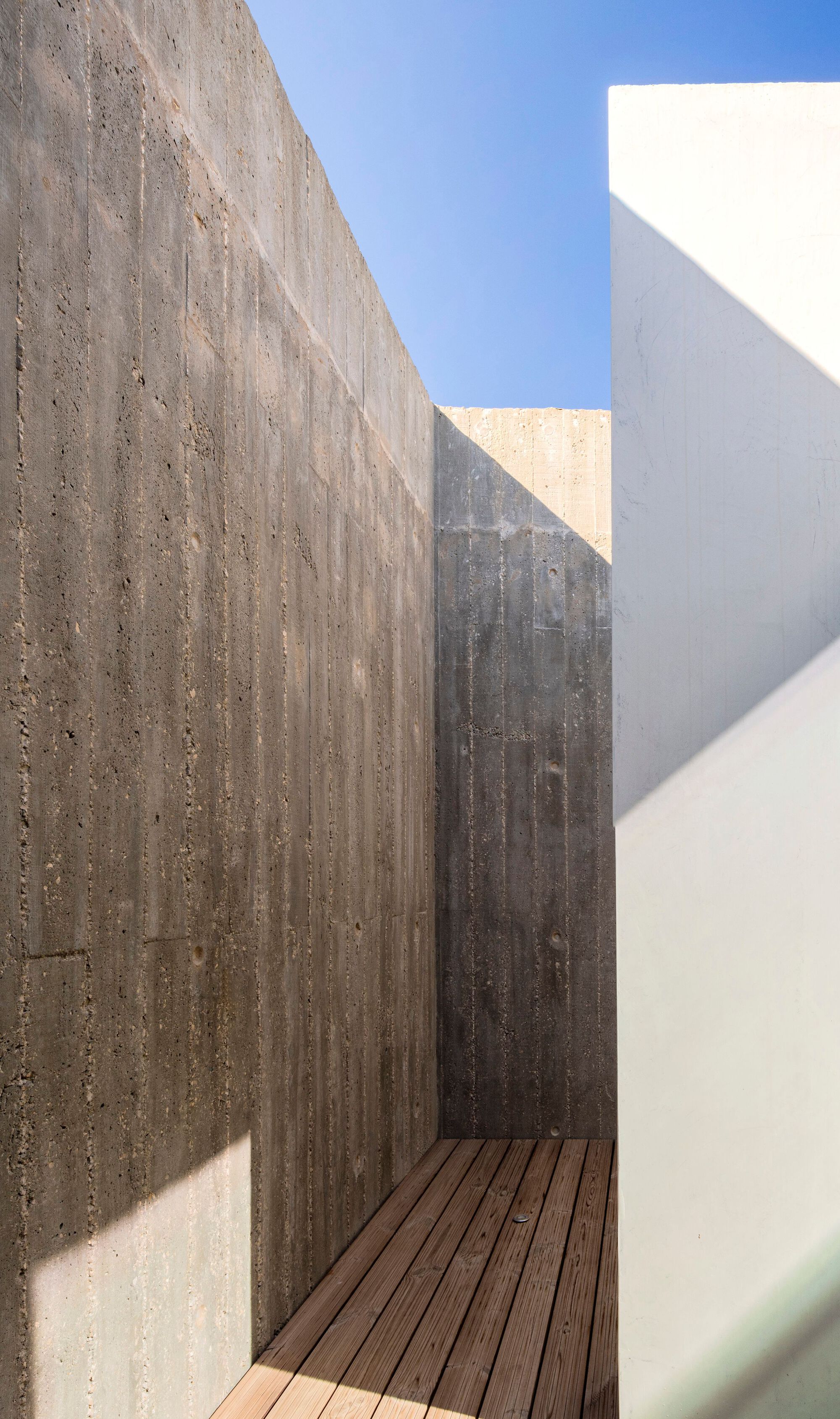 Narrow courtyard passage with board-formed concrete walls, timber decking and sharp afternoon shadows under blue sky