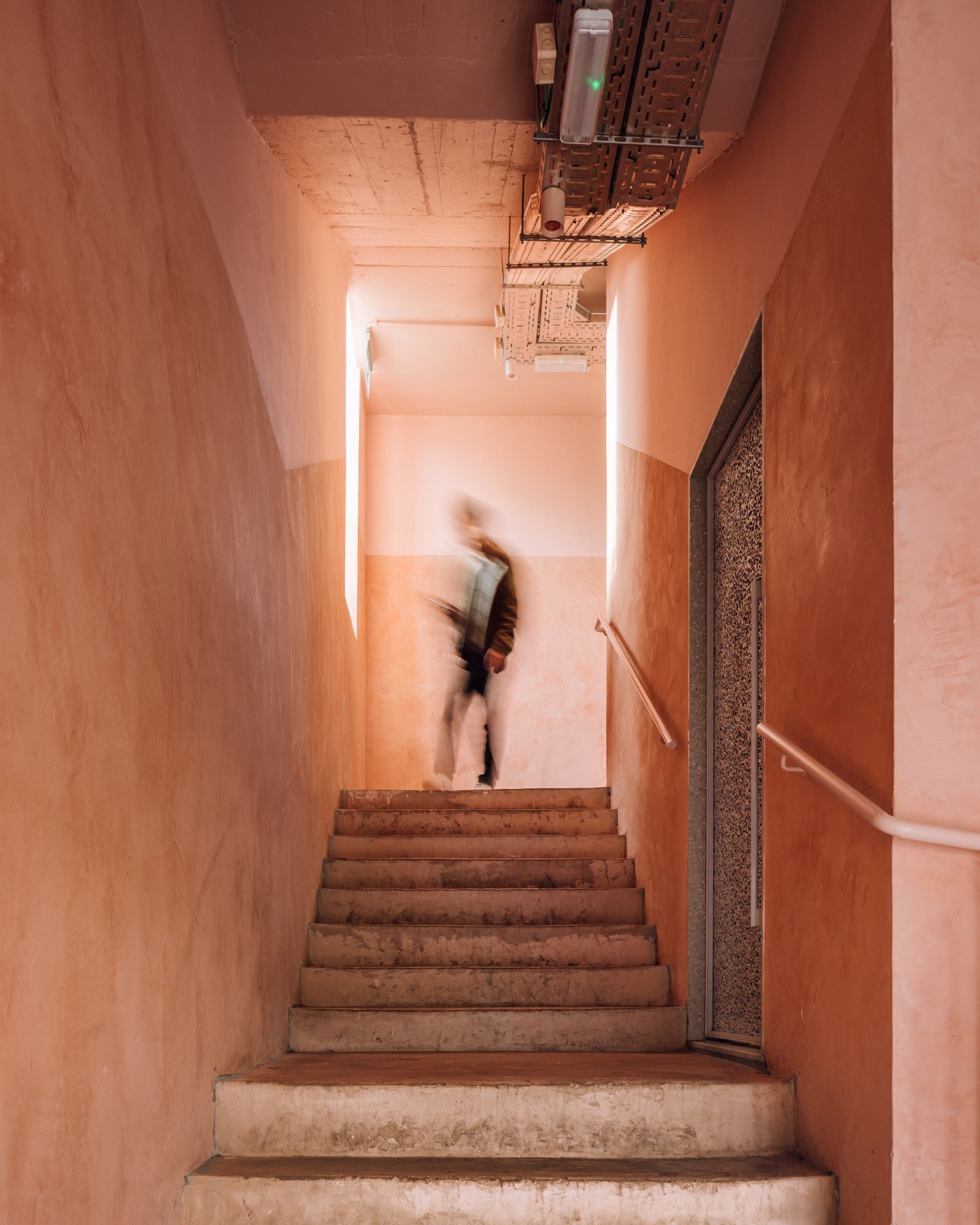 Narrow concrete staircase with pink plaster walls and a person in motion descending
