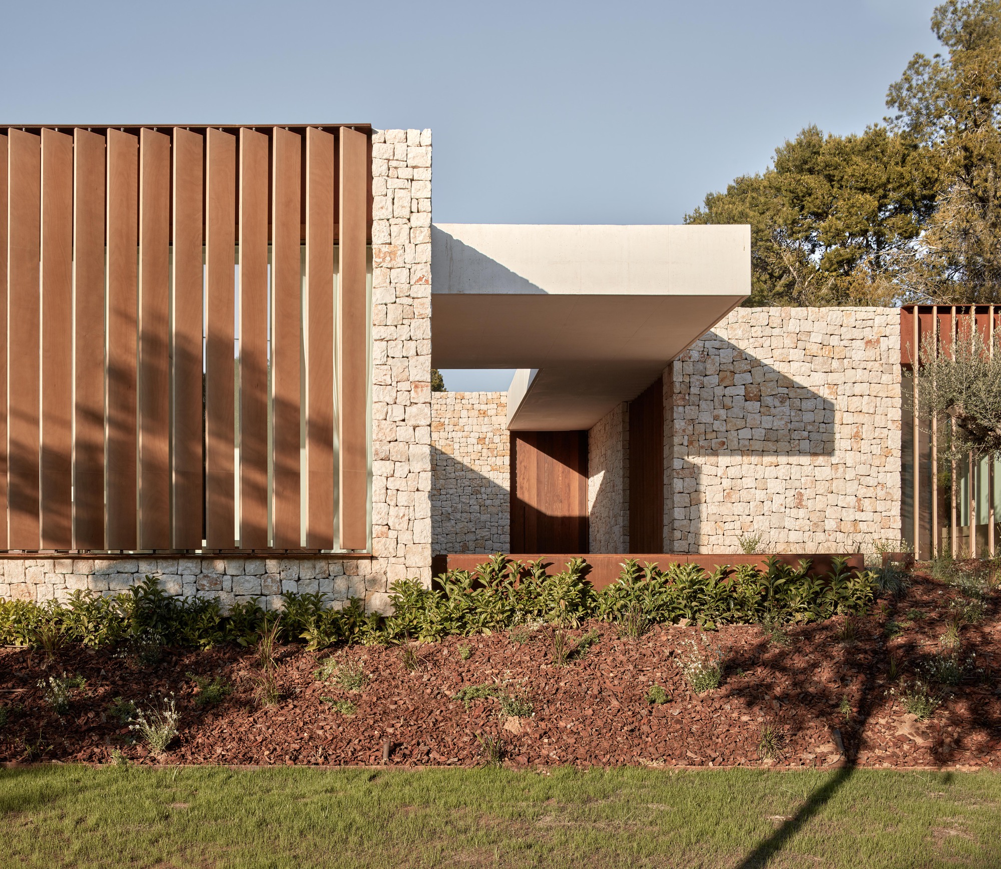Street facade with vertical corten steel panels, stone masonry, and white stucco volumes amid planted beds