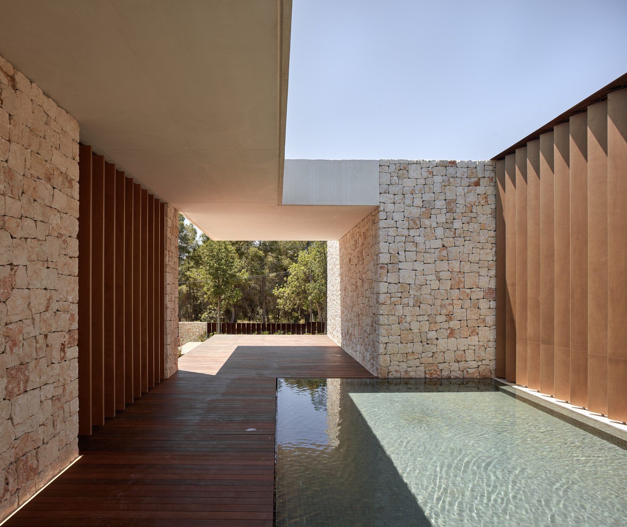 Covered passage with timber decking alongside reflecting pool flanked by stone walls and vertical metal screens