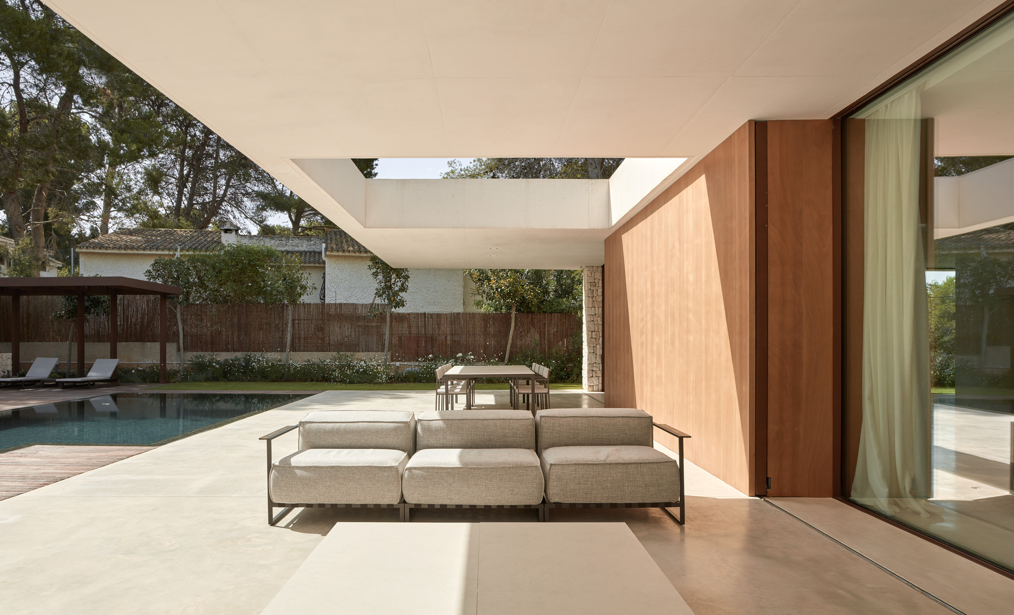 Covered outdoor terrace with wood-paneled pivot door and low sofa overlooking a pool and timber fence