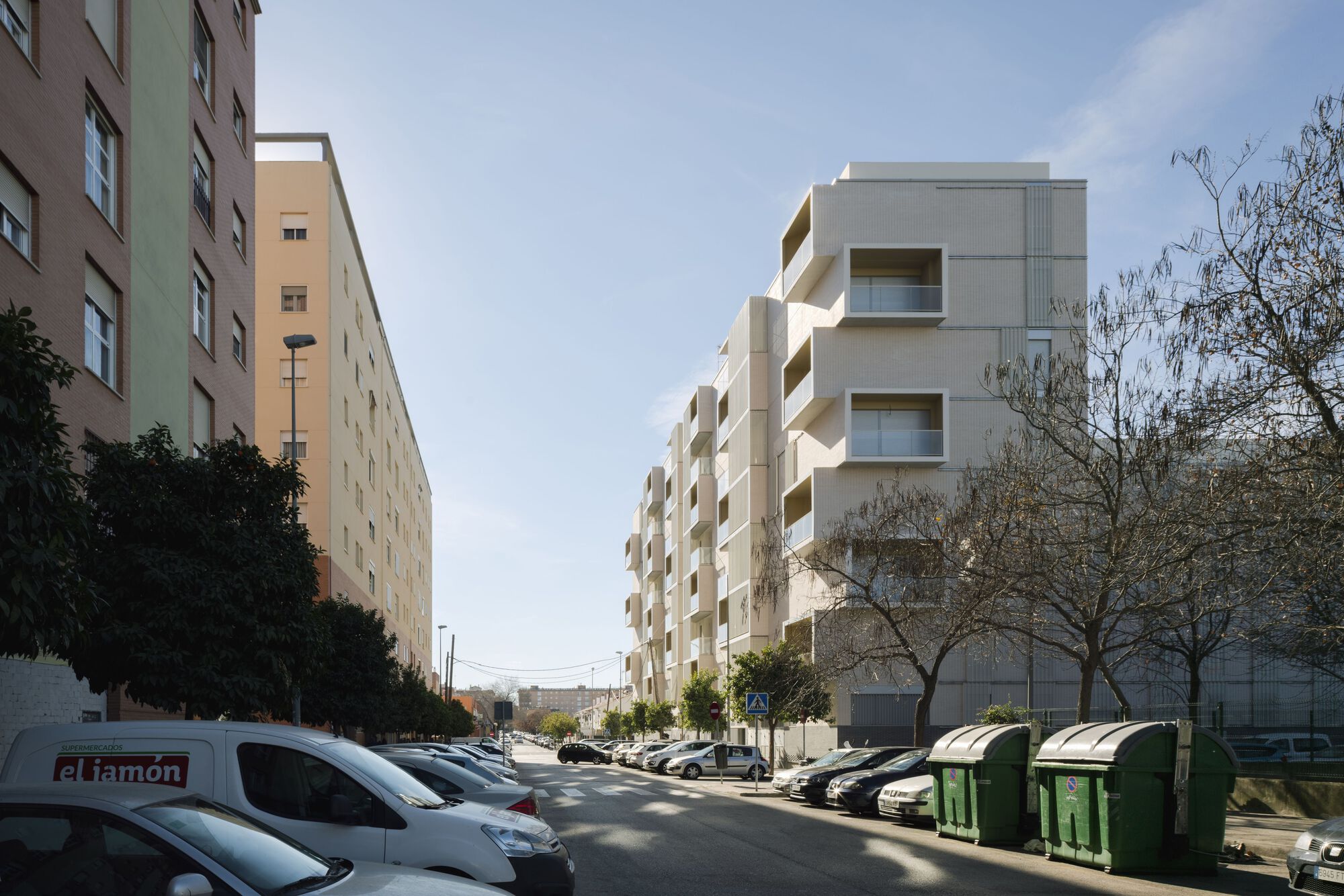 Street view of the stepped facade with recessed balconies, adjacent to parked cars and recycling bins