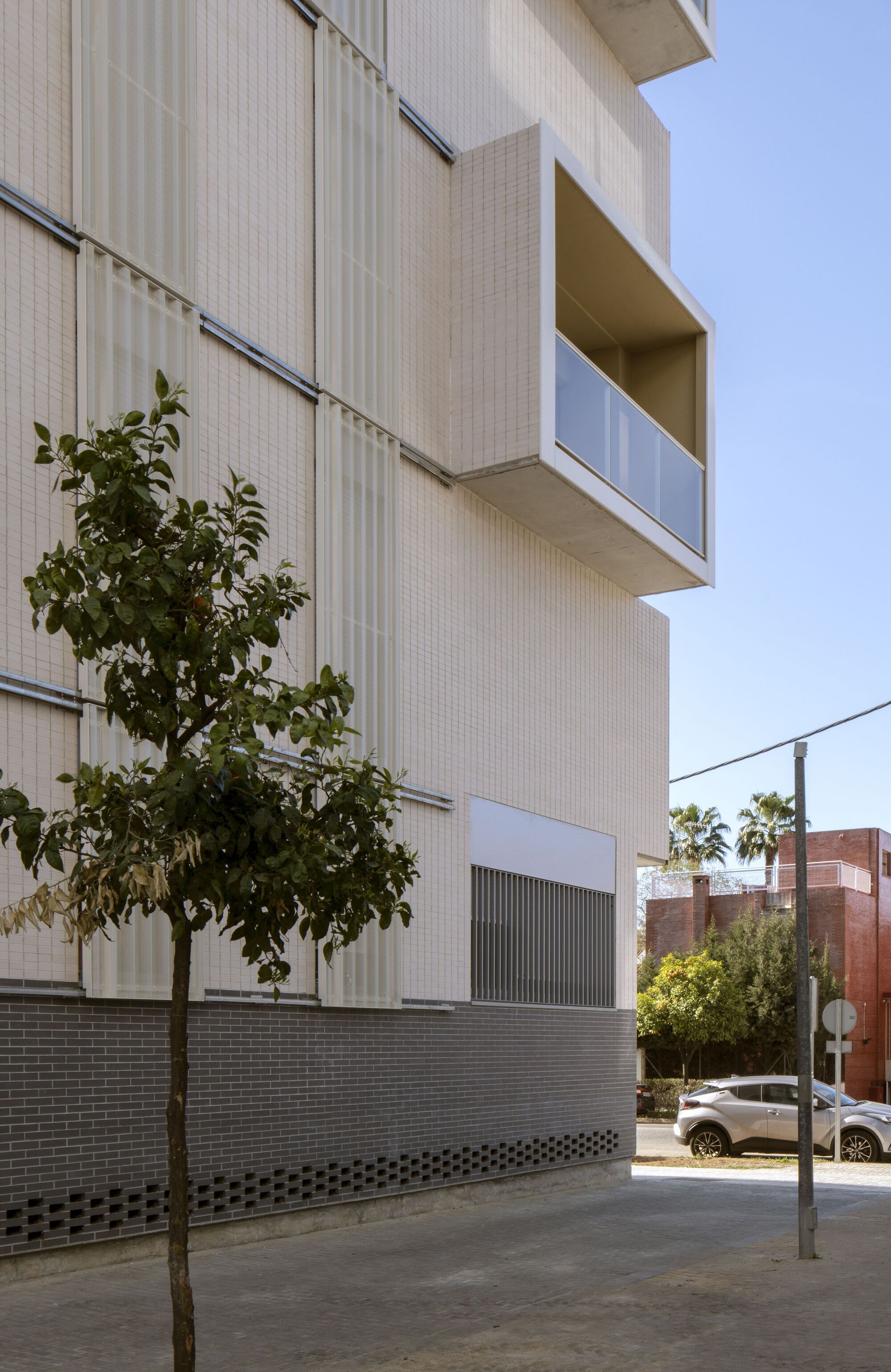 Close-up of the facade with white ribbed panels, recessed balcony, and street tree in sunlight