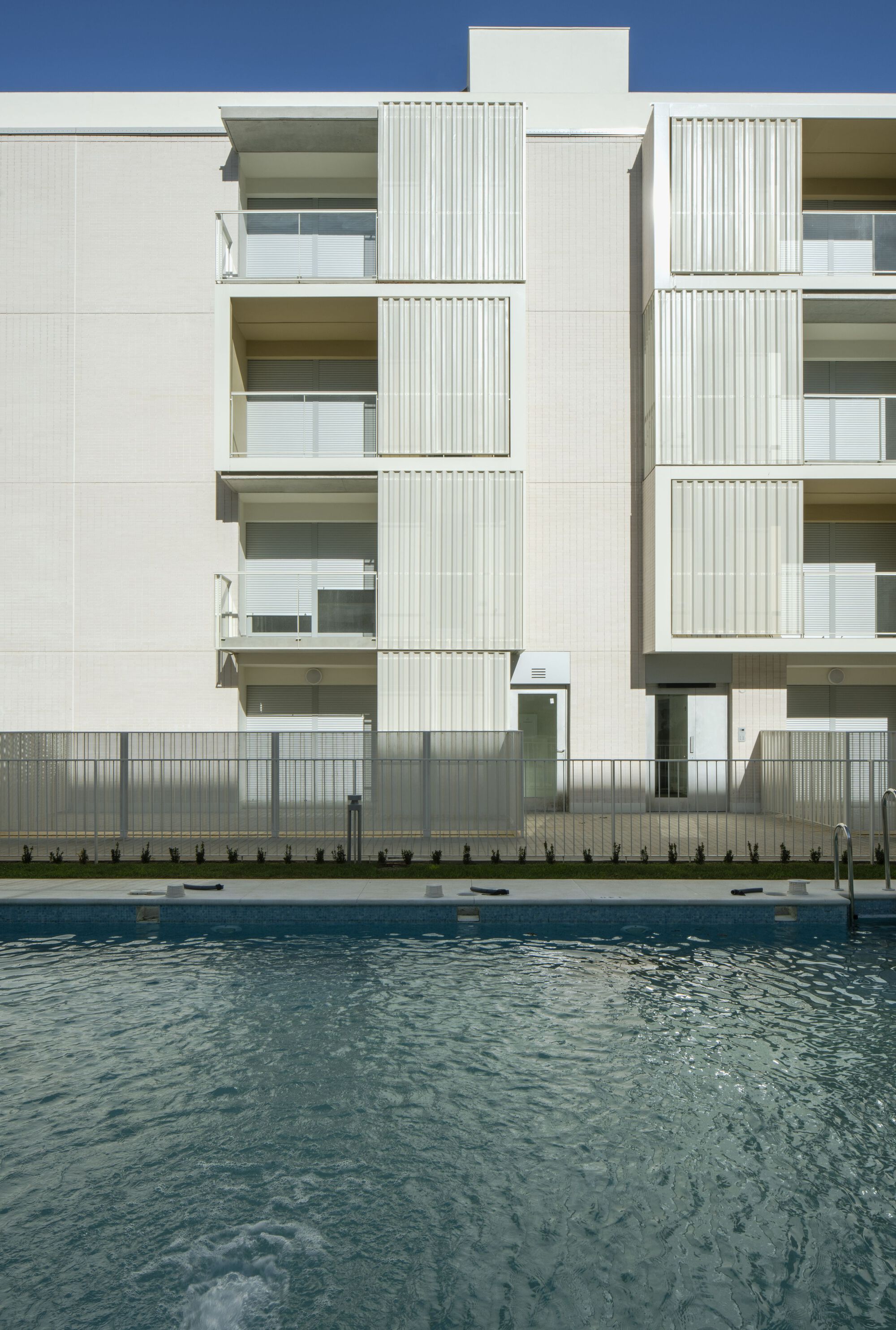 Poolside view of the white facade with recessed balconies and vertical louvers in clear daylight