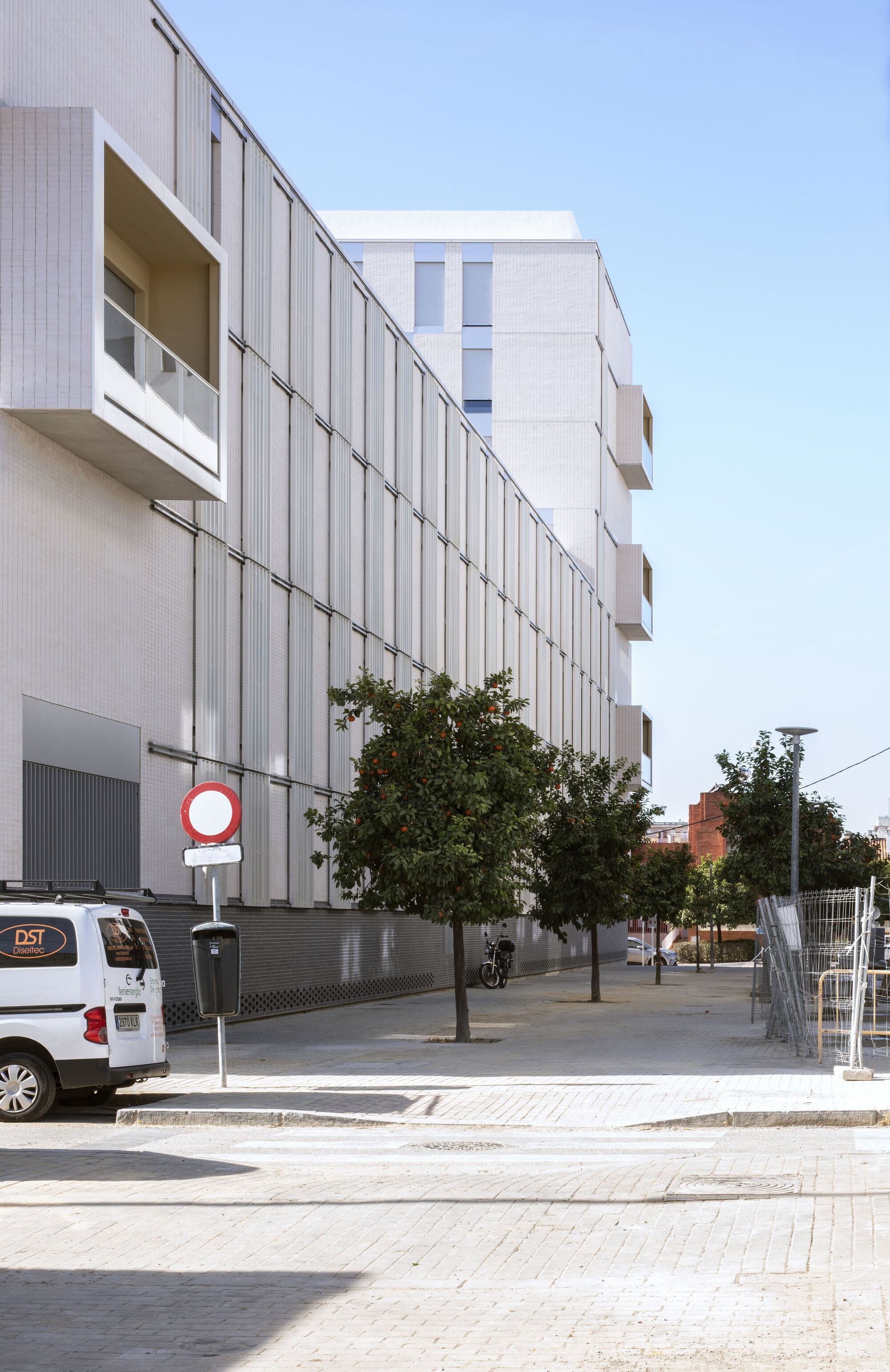 Long facade with vertical metal screening and street trees along a pedestrian sidewalk at dusk