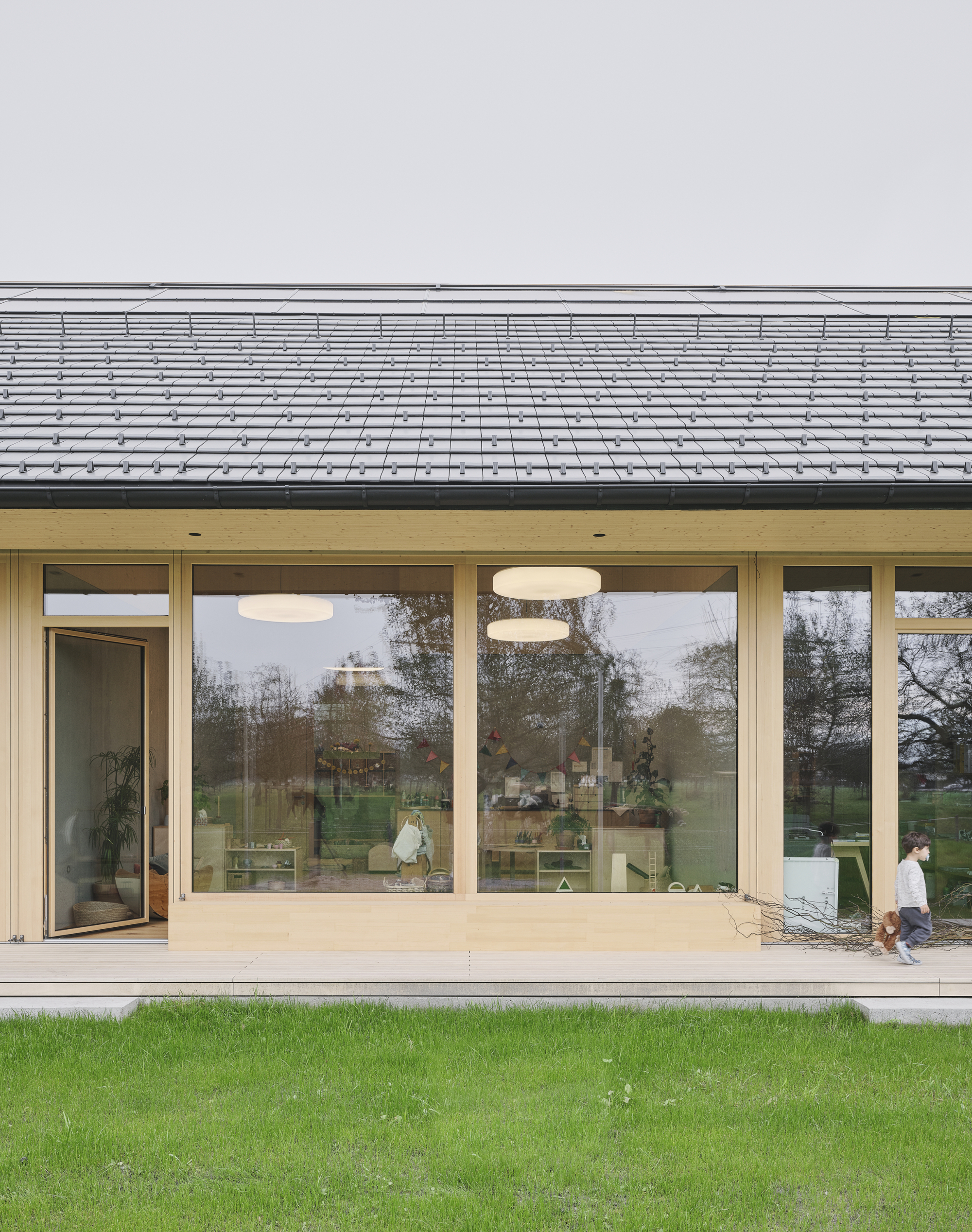 Glazed facade with timber frames beneath deep eave overhang and grey tile roof with child walking
